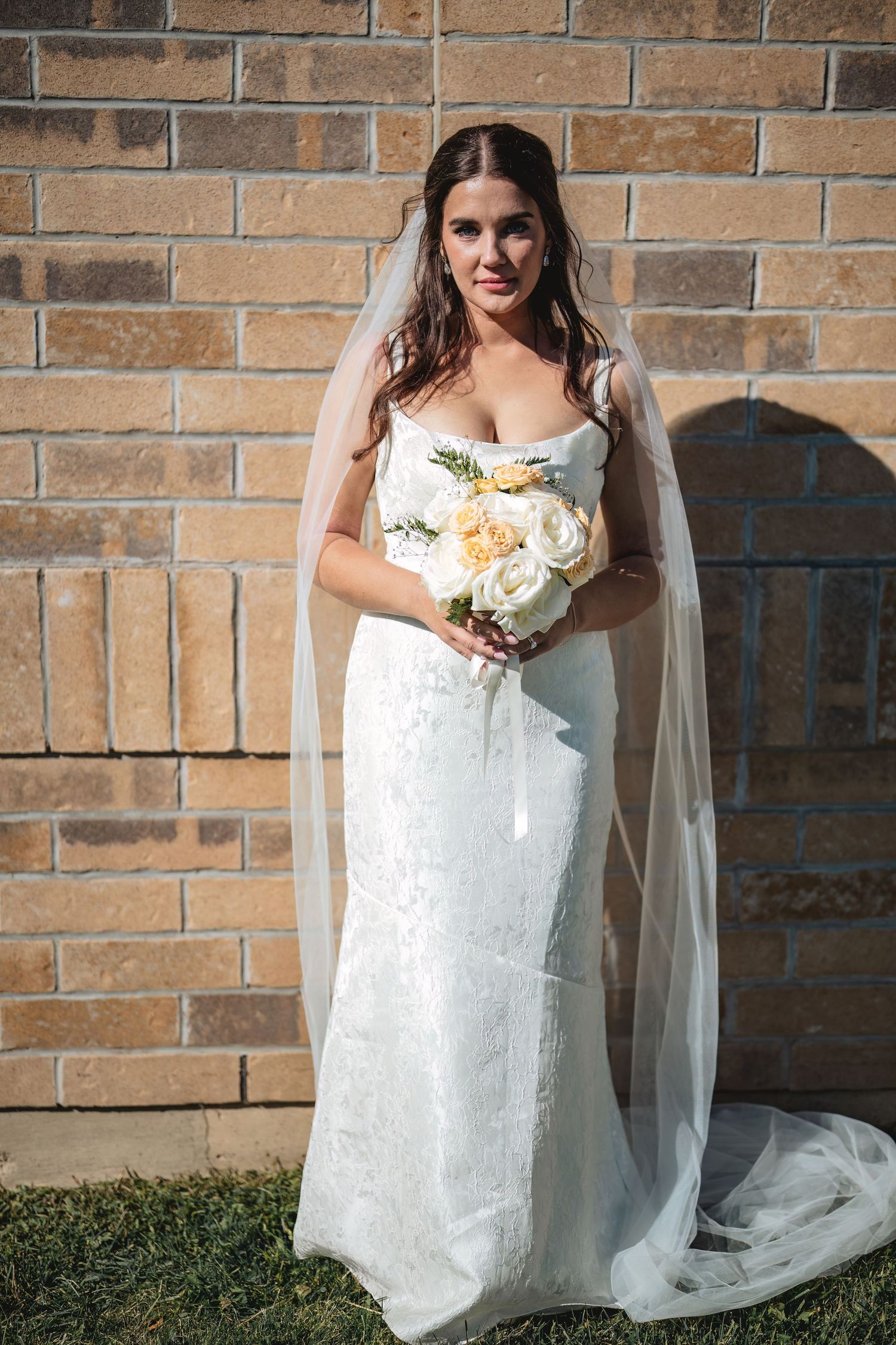 A bride in a fitted lace gown and veil holds a white bouquet against a brick wall backdrop.
