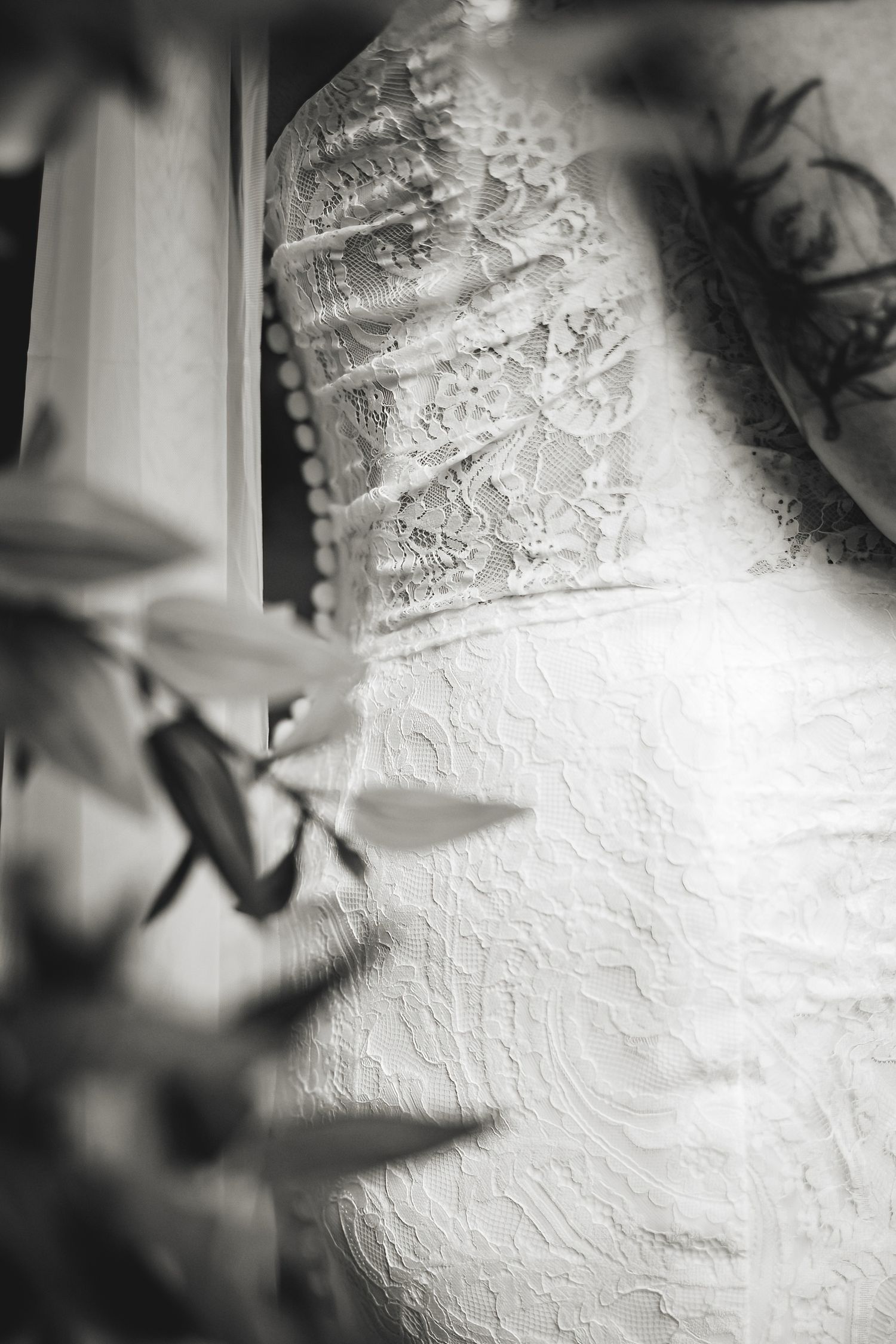 Black and white close-up of an elegant lace wedding dress hanging by a window with soft natural light.