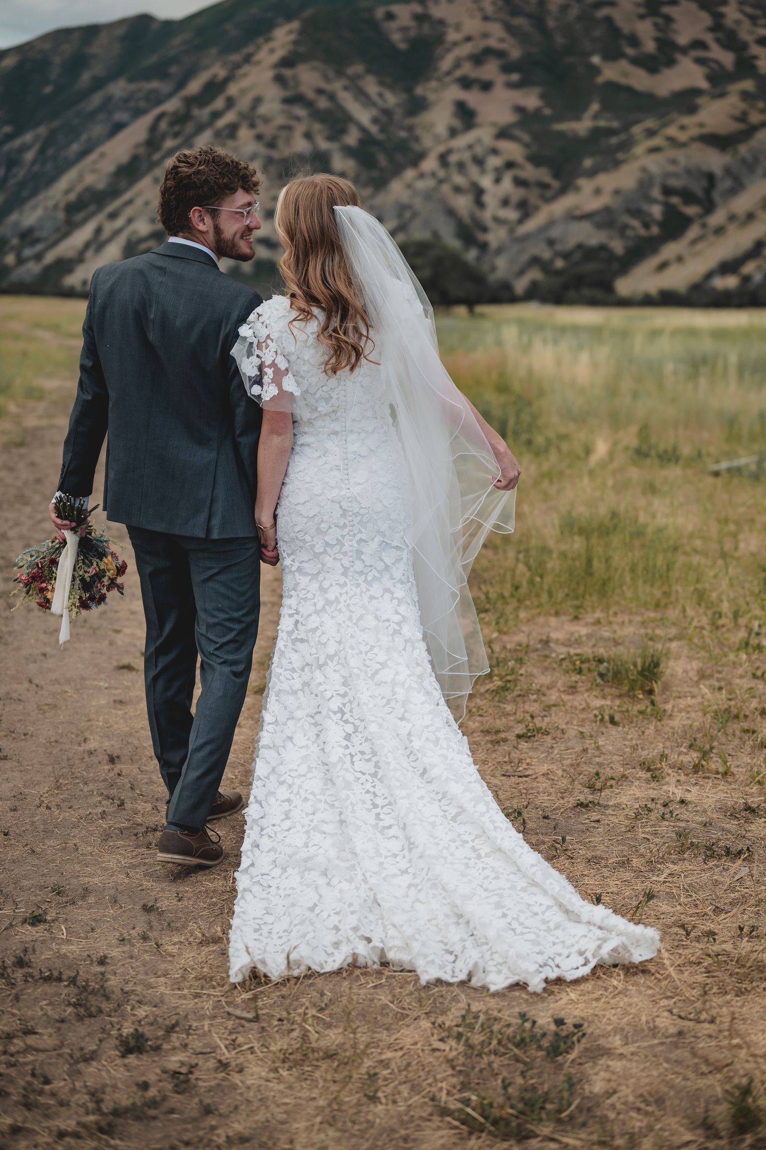 Bride in lace wedding gown with veil walks hand-in-hand with groom through mountain field.