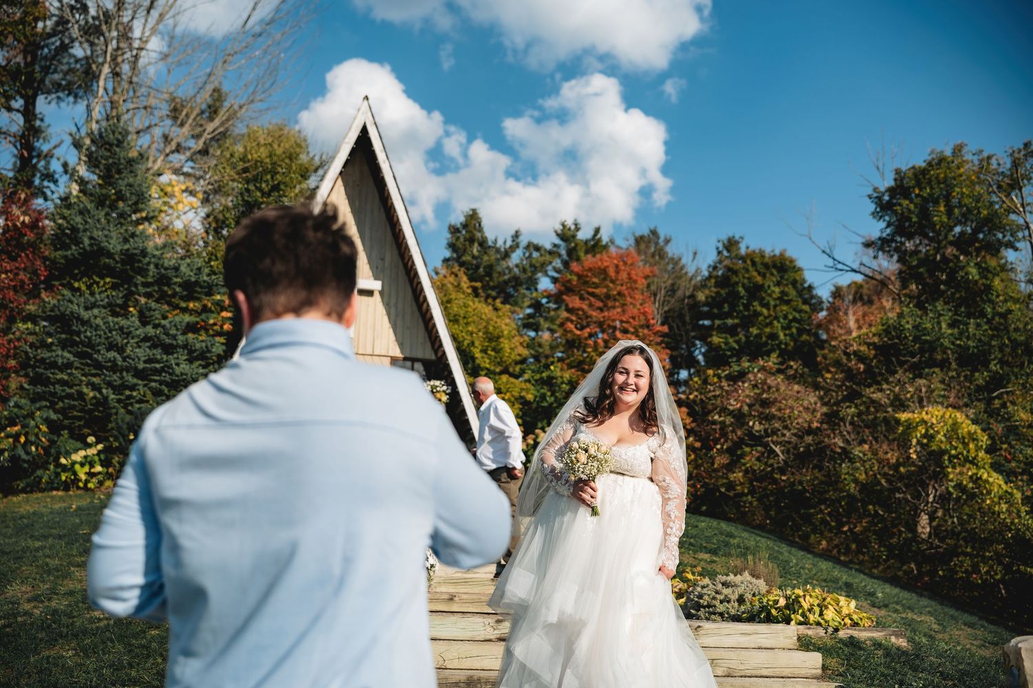 Bride in white gown smiles holding bouquet approaching groomsman outside a chapel outdoors.