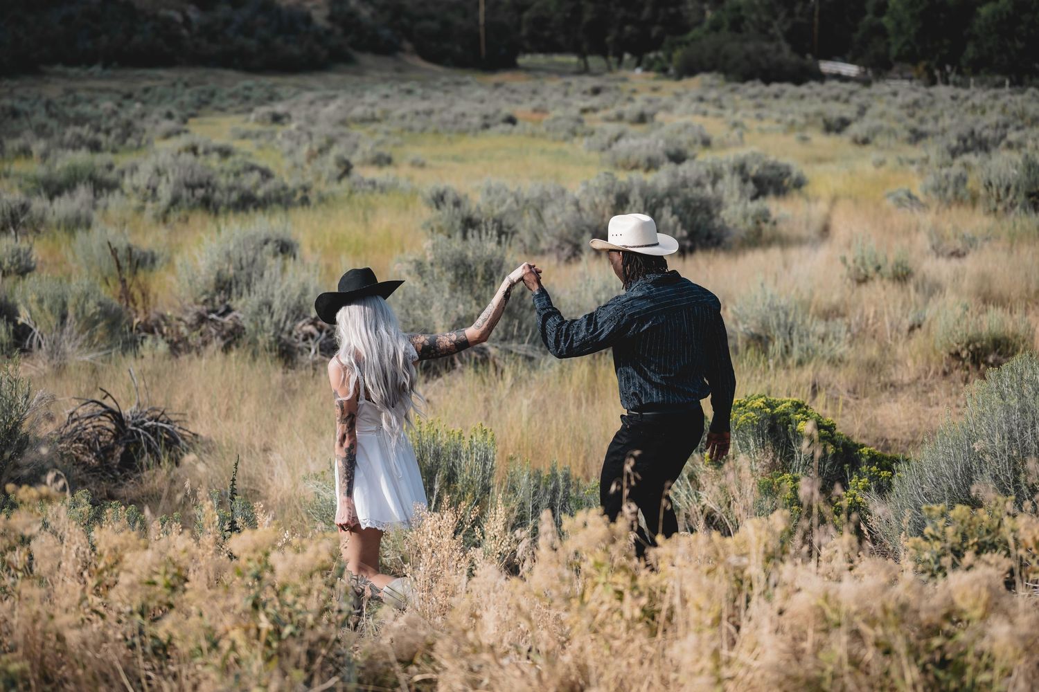 Couple in cowboy hats dance romantically in golden sagebrush field during outdoor engagement session.