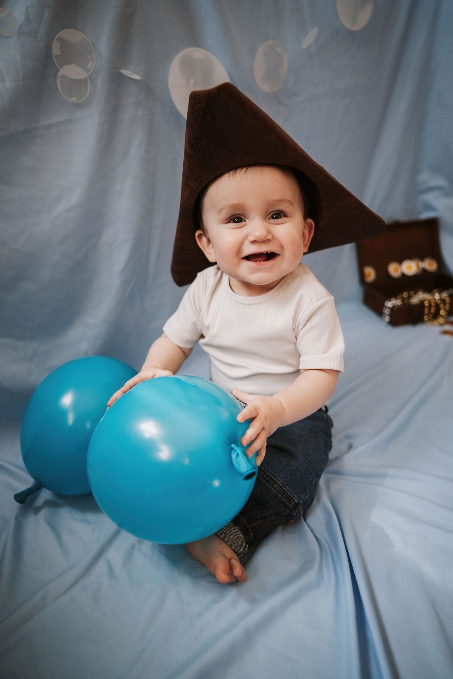 A smiling baby wearing a pirate hat sits on a blue blanket holding two blue balloons.