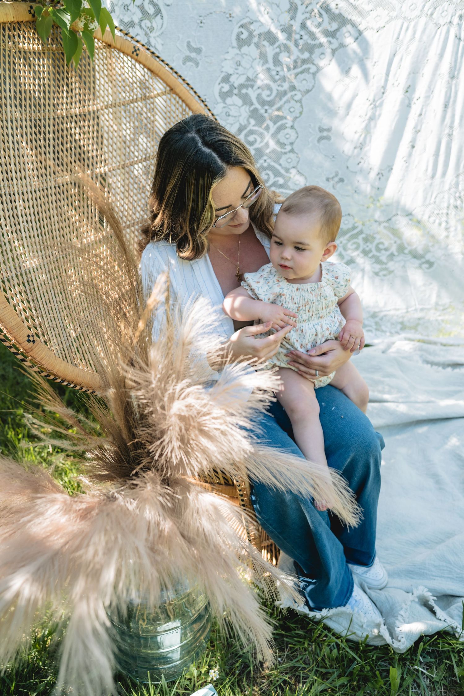 Mother holding baby in boho outdoor setting with peacock chair, pampas grass, and lace backdrop in natural sunlight.