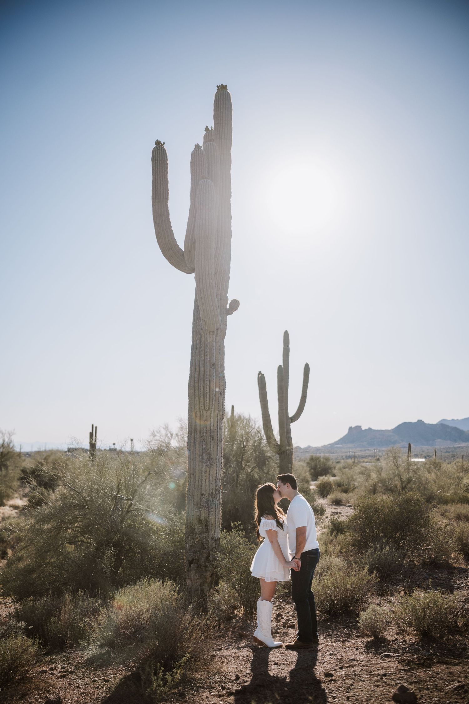 Couple kisses holding hands beside tall saguaro cactus in sunny Arizona desert landscape.
