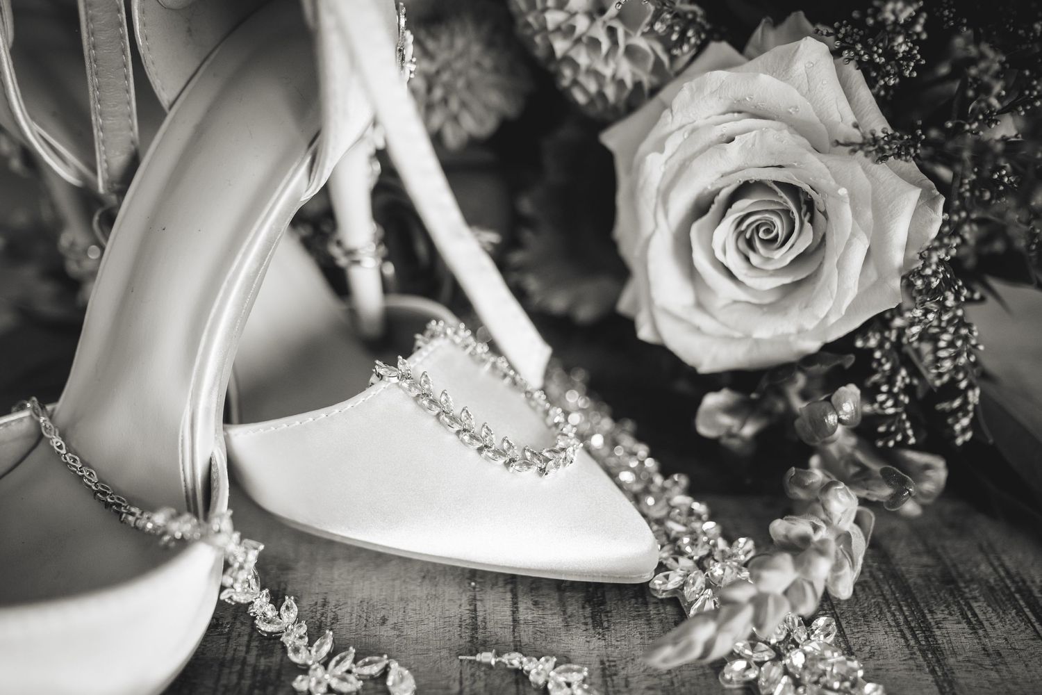 Black and white close-up of white bridal heels with crystal buckles beside a rose bouquet.