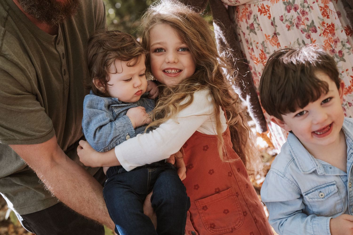 A young girl hugs a toddler while a smiling boy looks on during a warm family outdoor session.