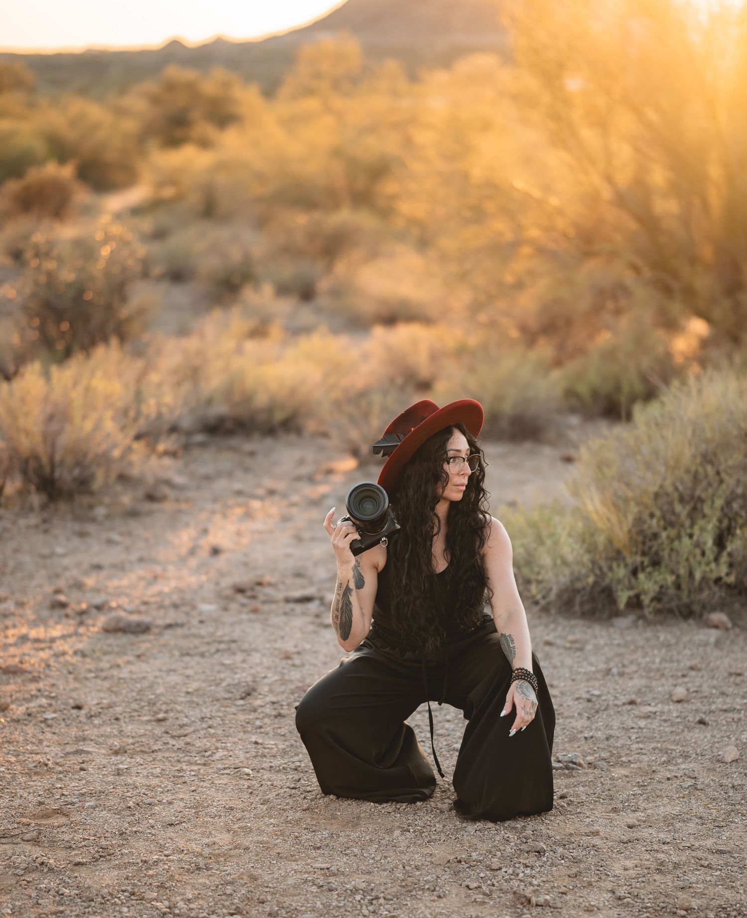 Woman in red hat and black outfit kneeling in desert at golden hour holding a camera.