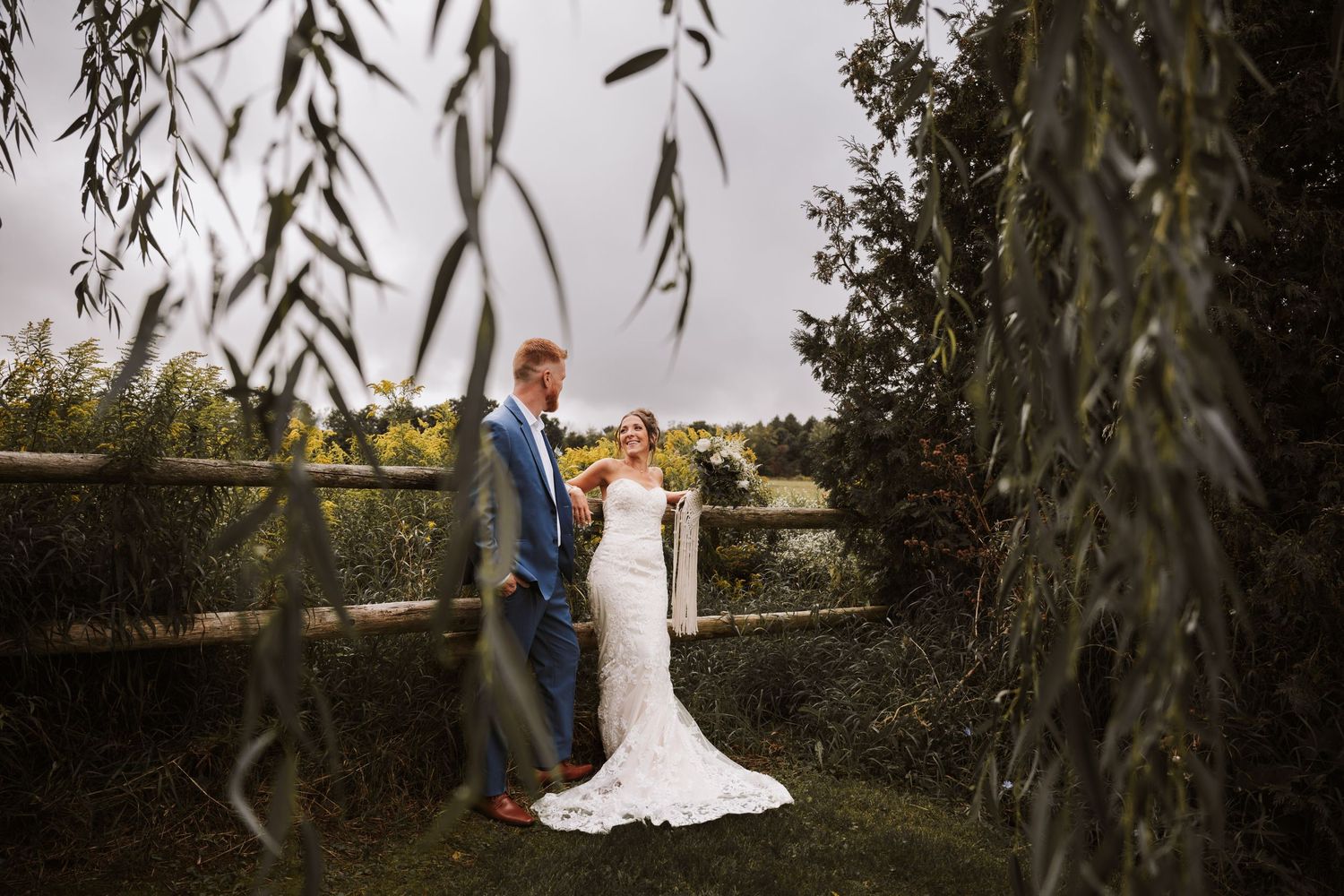 A bride in a lace gown and groom in a blue suit stand romantically by a wooden fence, framed by weeping willow branches.