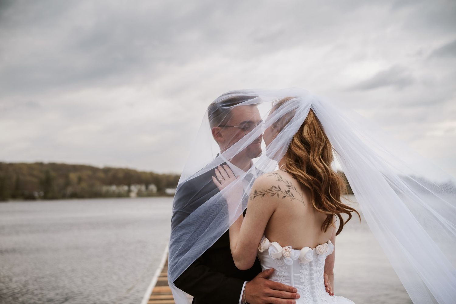 Bride and groom share a romantic kiss under a flowing white veil on a lakeside dock on their wedding day.