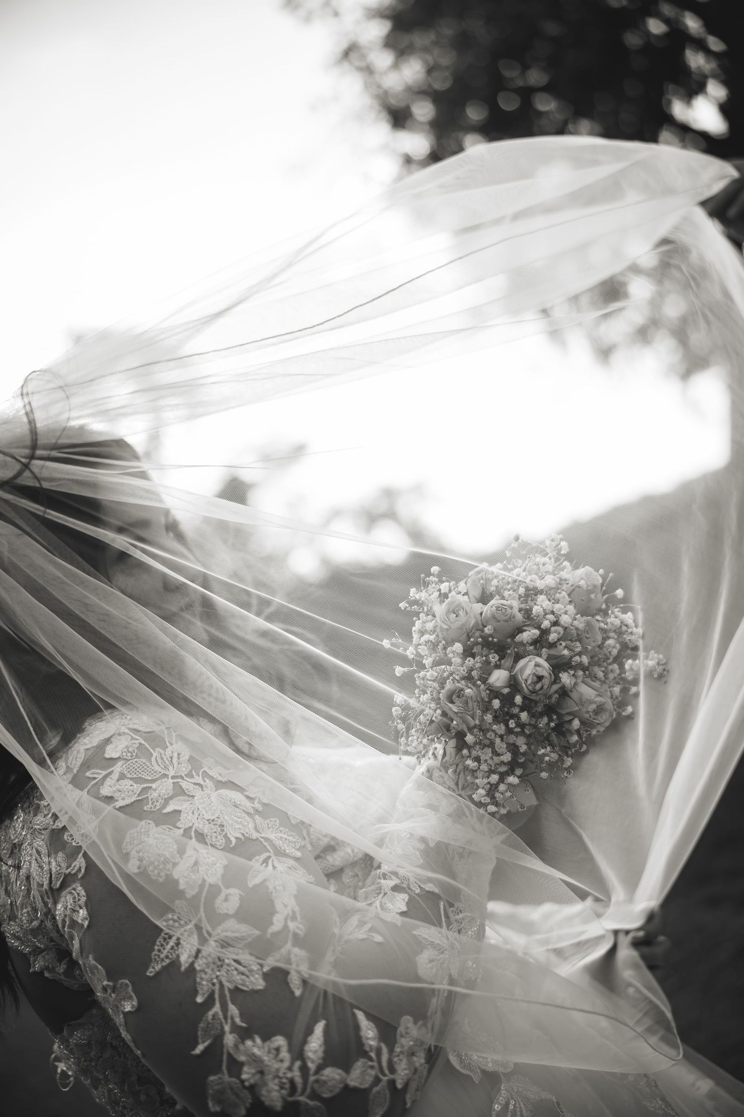 Black and white image of bride with veil blowing in wind, holding floral bouquet.