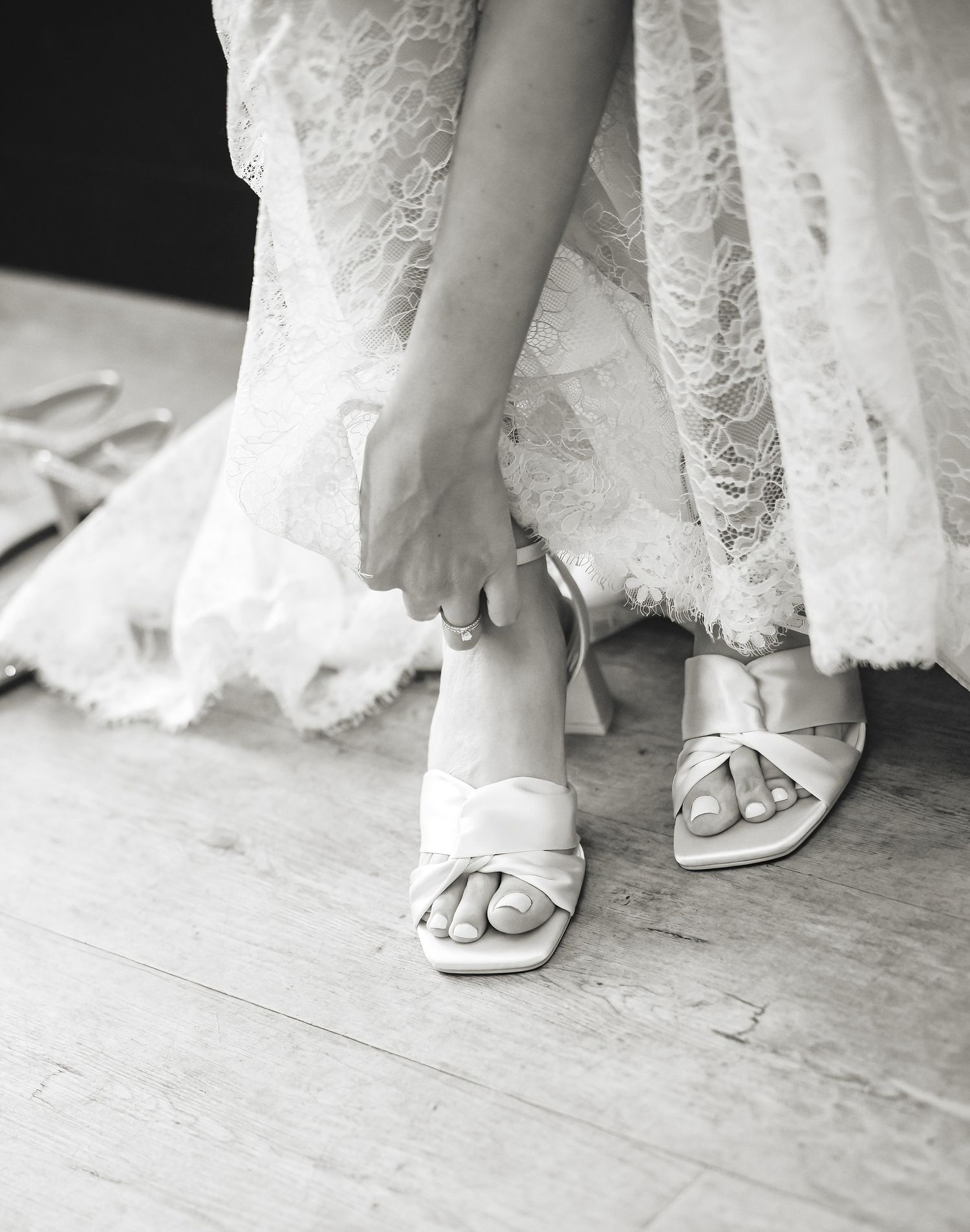 Bride in lace wedding dress adjusting white strappy heeled sandals on wooden floor, black and white photo.