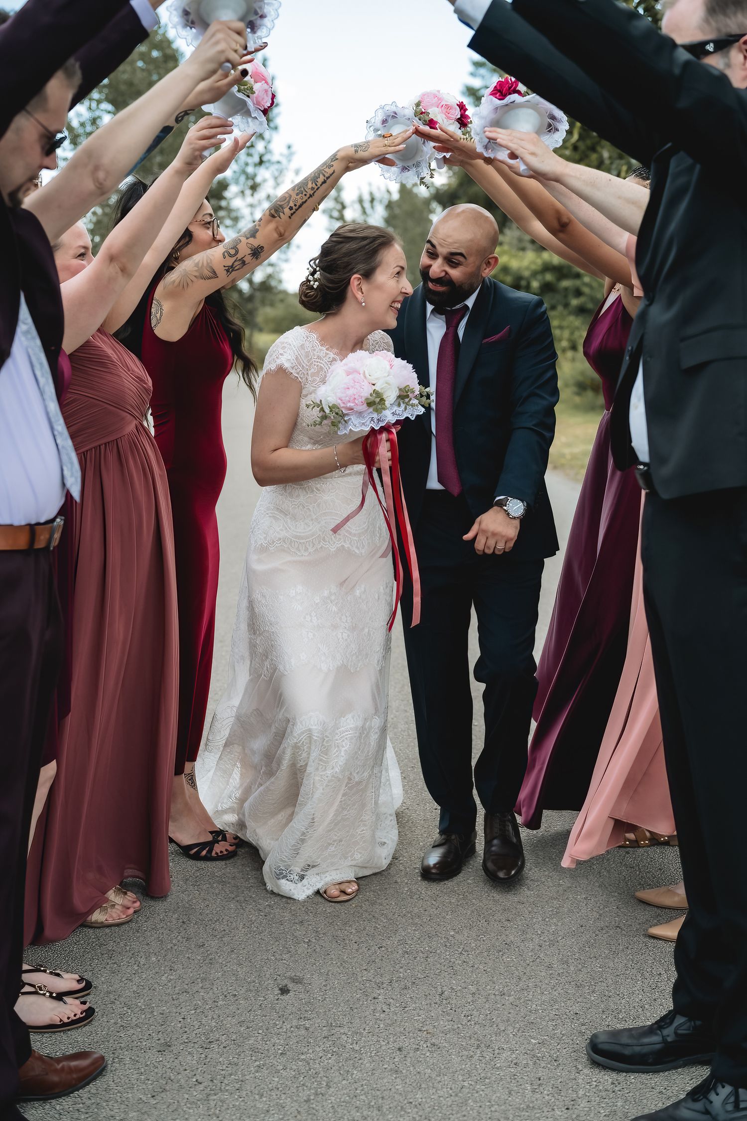 Bride and groom walk through arch of raised bouquets held by wedding party in burgundy and mauve gowns outdoors.