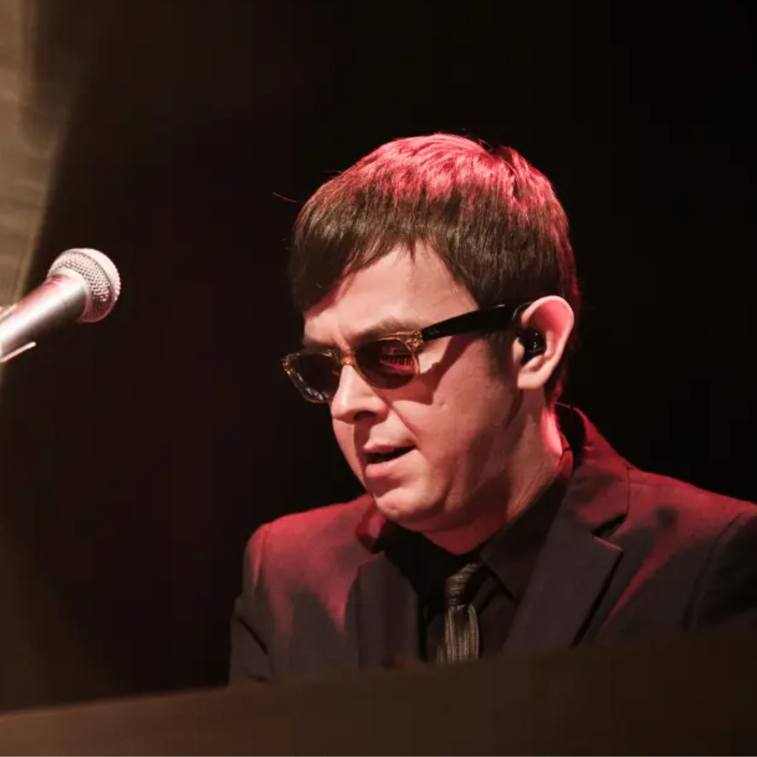 A performer in dark glasses sings into a microphone on a dimly lit stage with dramatic red lighting.