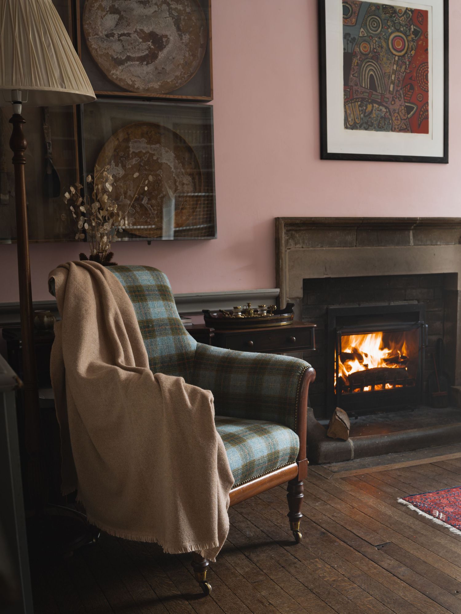 A cozy armchair with plaid upholstery sits beside a lit fireplace in a room with pink walls. Blanket product shoot for Ava Innes.
