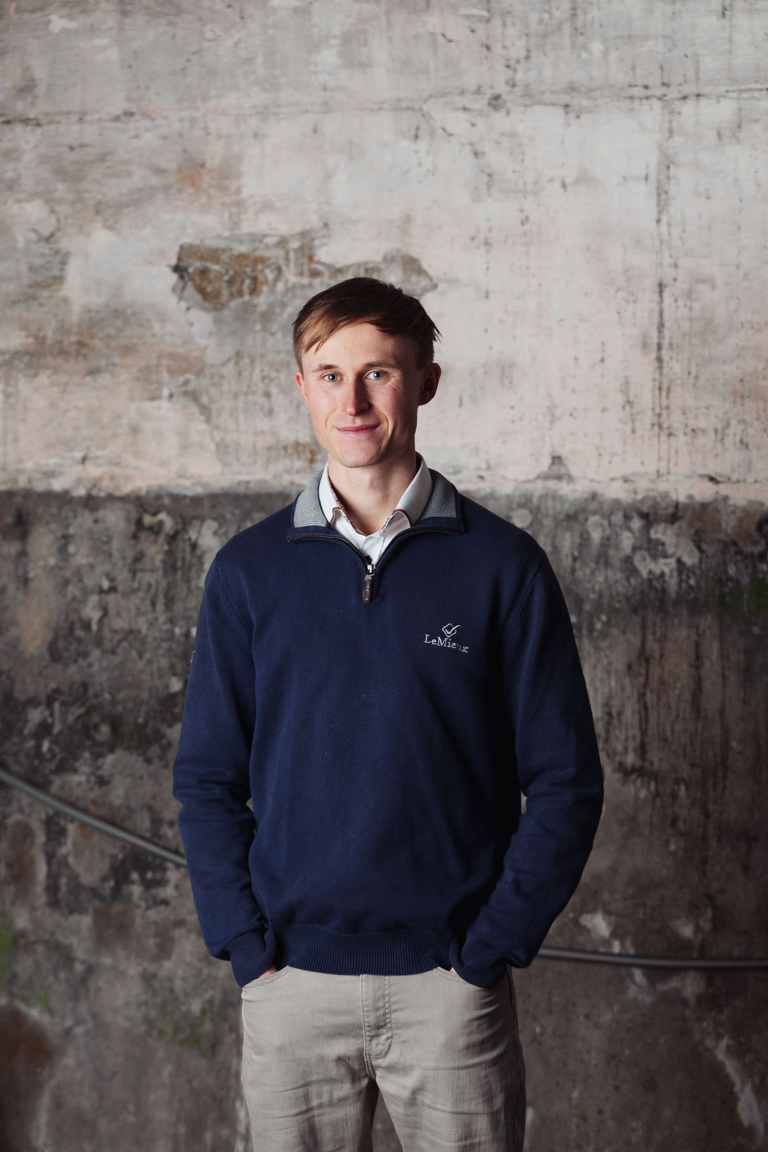 Professional portrait against industrial concrete wall wearing navy sweater and light pants. Lewis McLean.