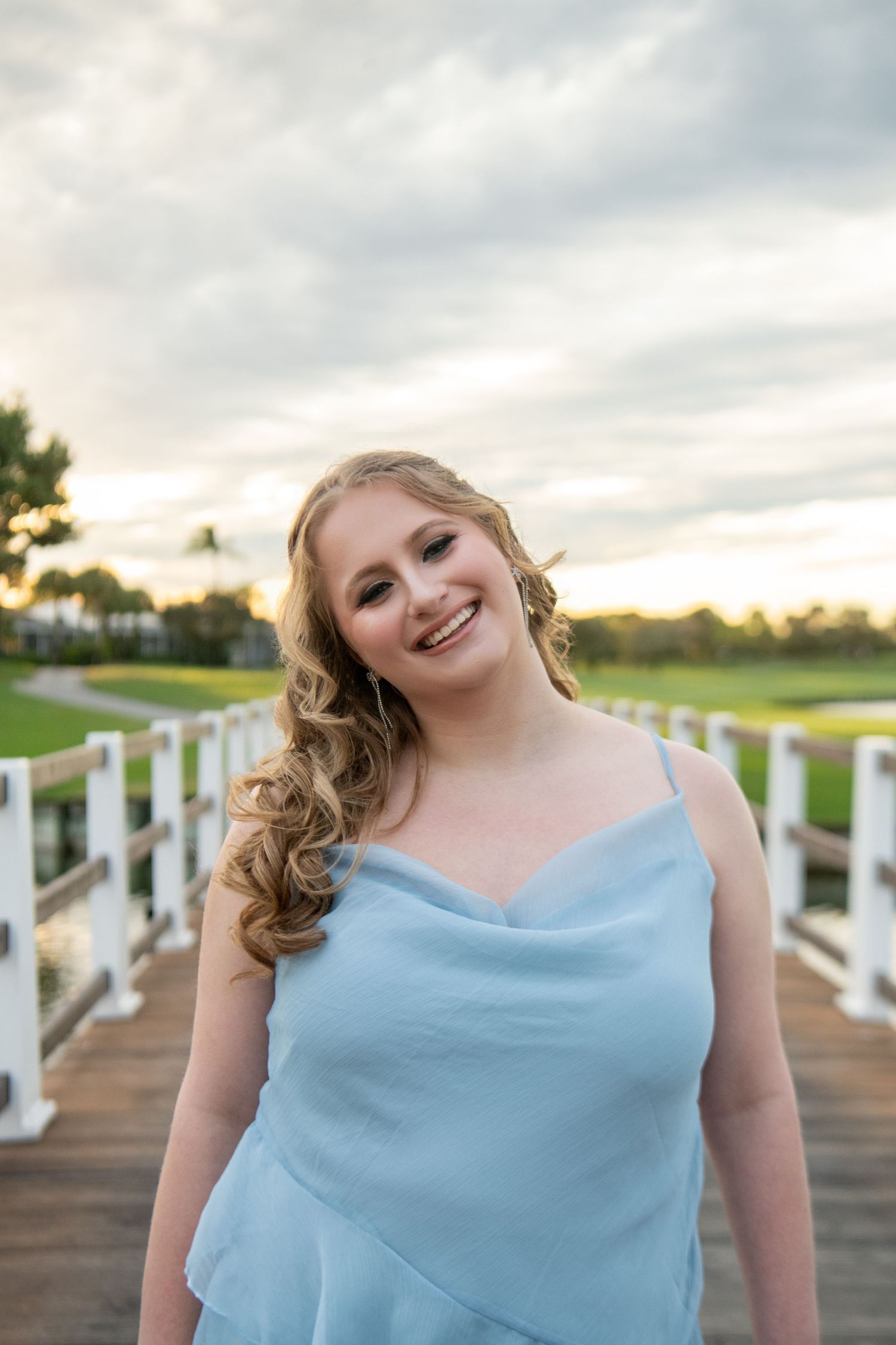 A woman in a light blue dress smiles while standing on a wooden boardwalk at sunset.