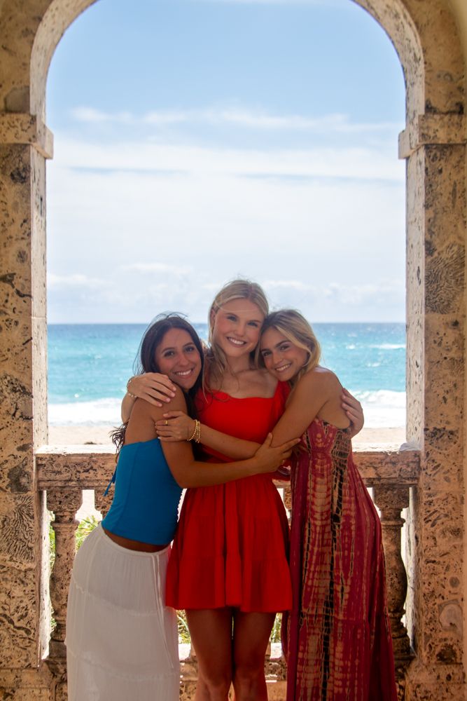 Group enjoying a sunny day at a beachfront stone colonnade with turquoise ocean waters visible in the background.