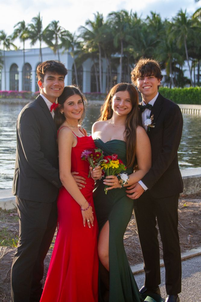 Two couples in formal attire pose together for prom photos near palm trees and waterfront at sunset.