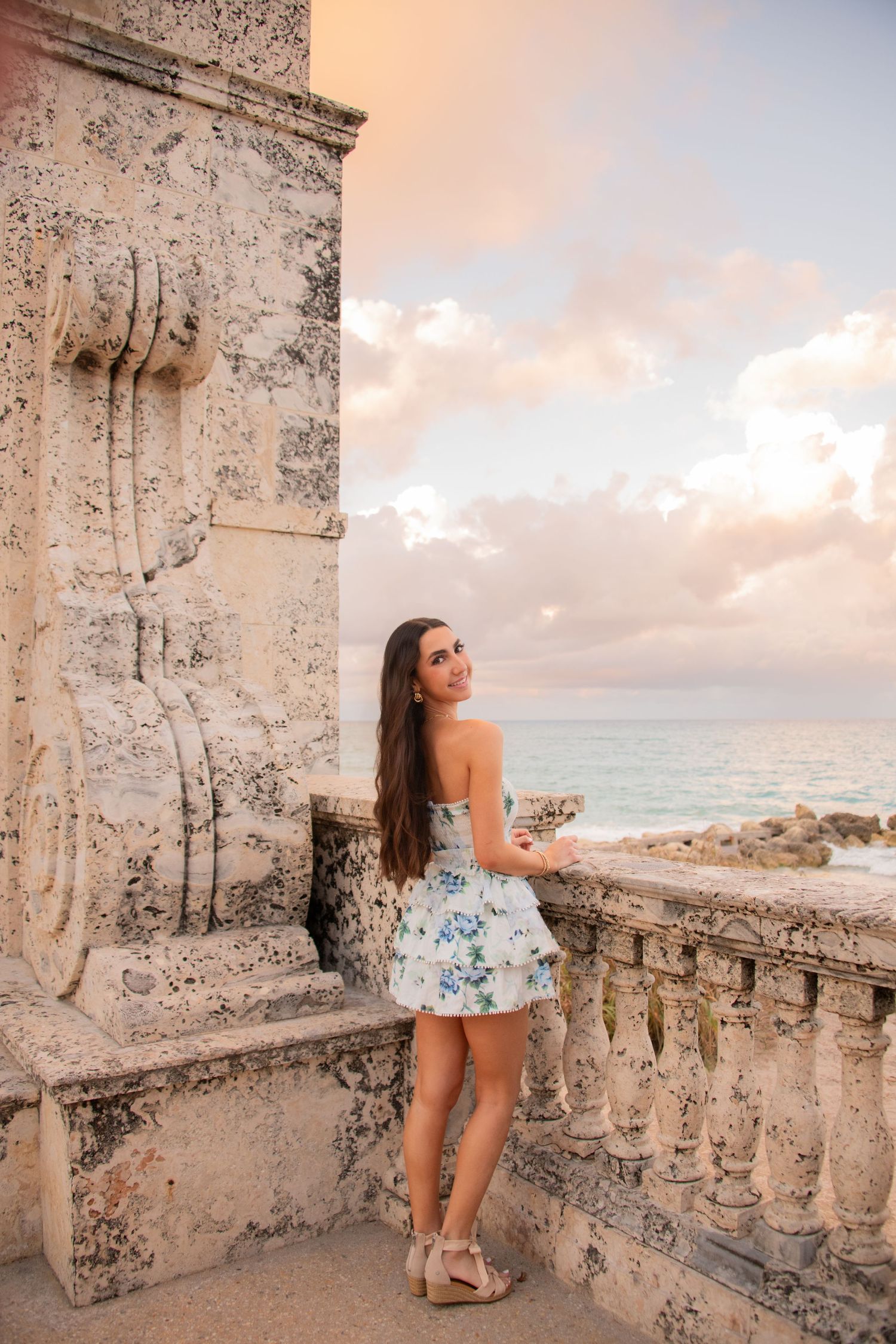 A person in summer attire enjoys sunset views from a stone balcony overlooking the ocean at a historic coastal location.