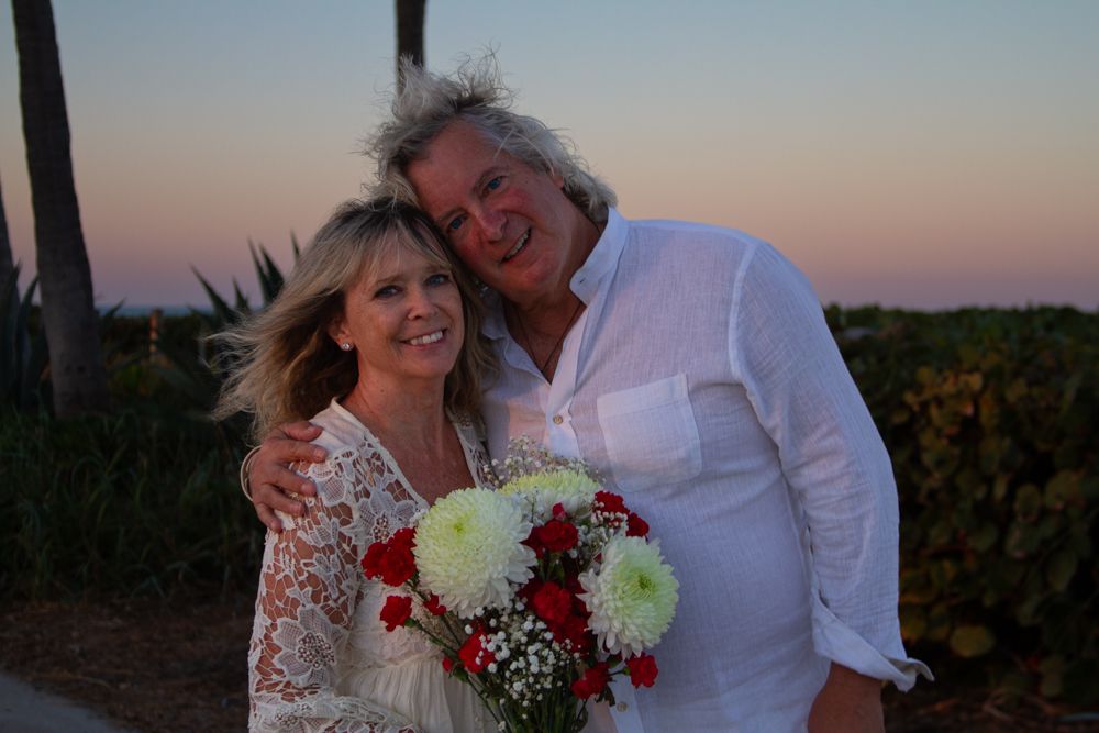 A couple in white attire poses together at sunset holding a bouquet of white and red flowers.