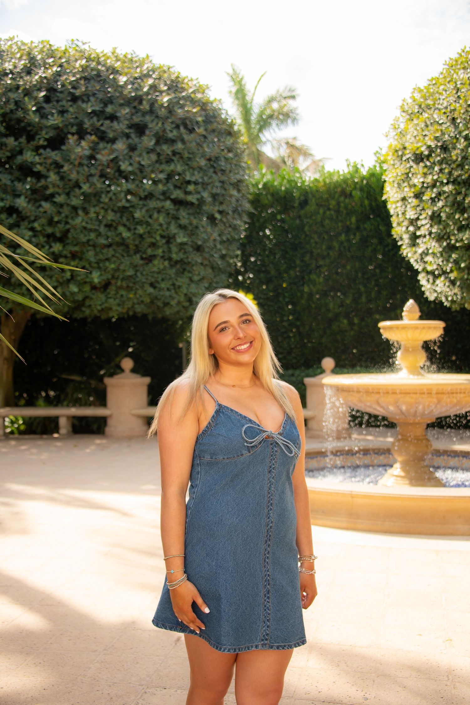 Woman in denim mini dress poses in garden with fountain and manicured hedges in background.