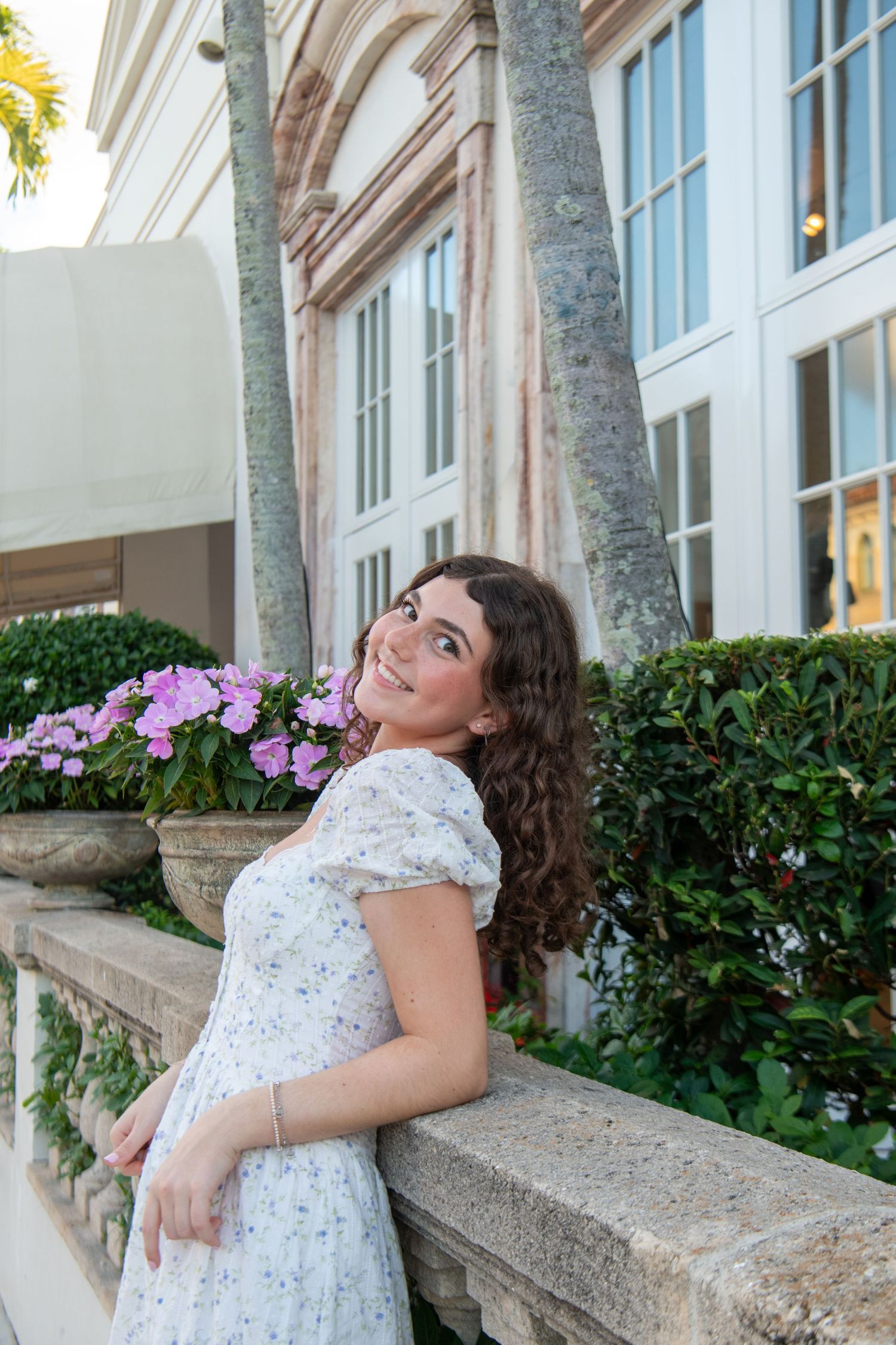 A person in a white floral dress poses playfully on a stone balcony with palm trees and windows in the background.