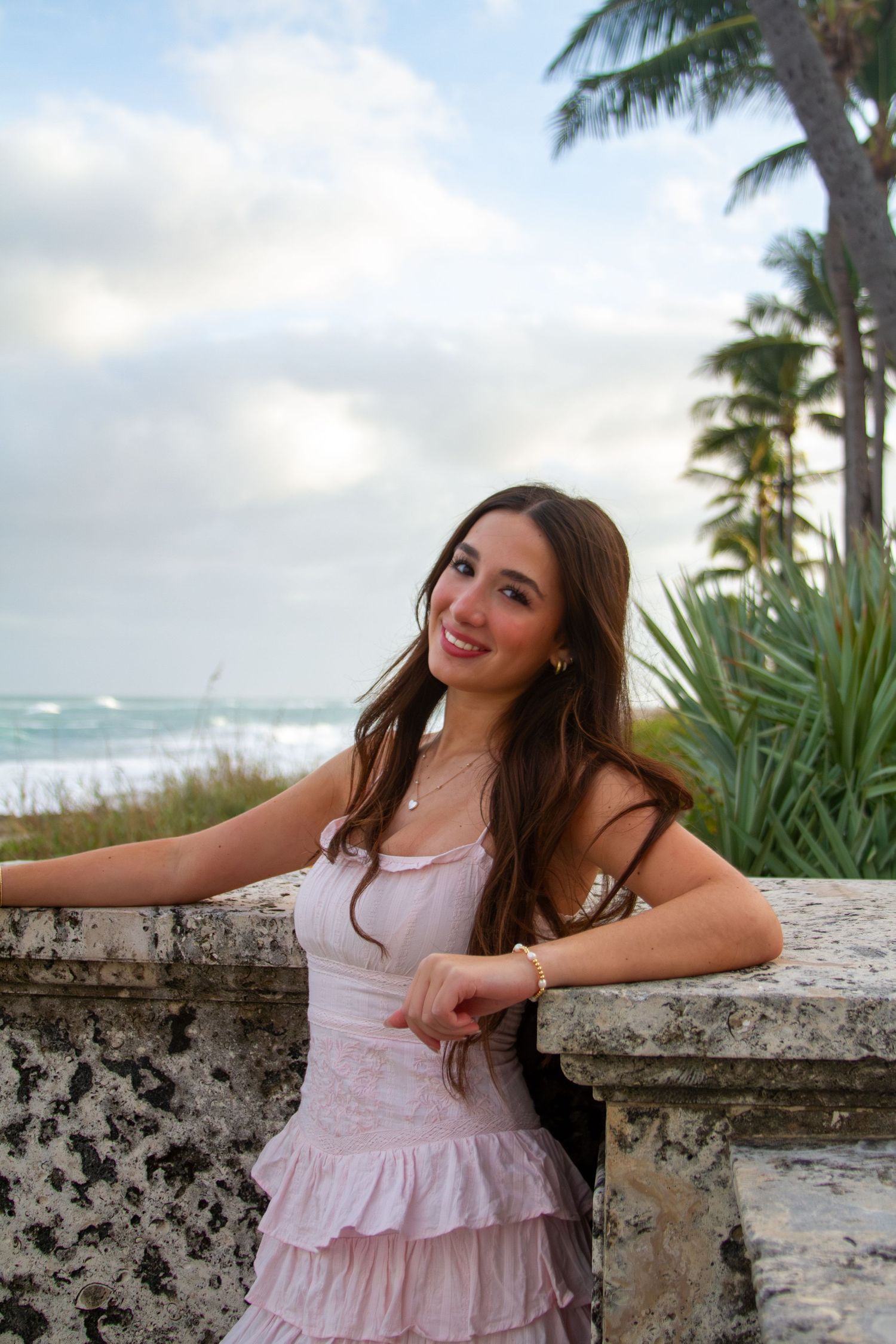 A person in a pink ruffled dress poses against a stone wall with palm trees and ocean waves in the background.