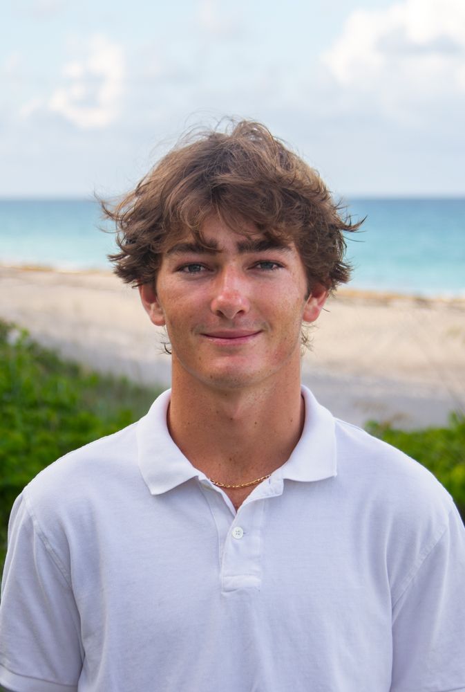 Young person in white polo shirt smiling at camera with tropical beach and turquoise ocean in background.