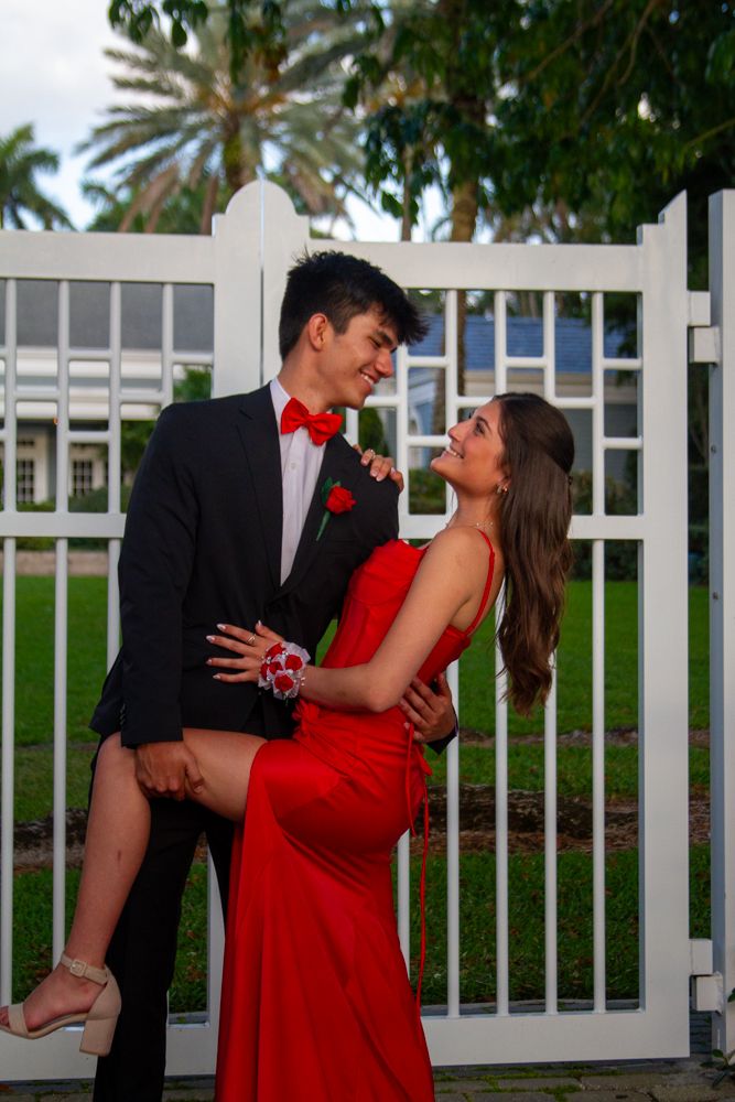 A couple in formal attire poses playfully by a white gate, with the woman in a red dress and man wearing a bowtie.