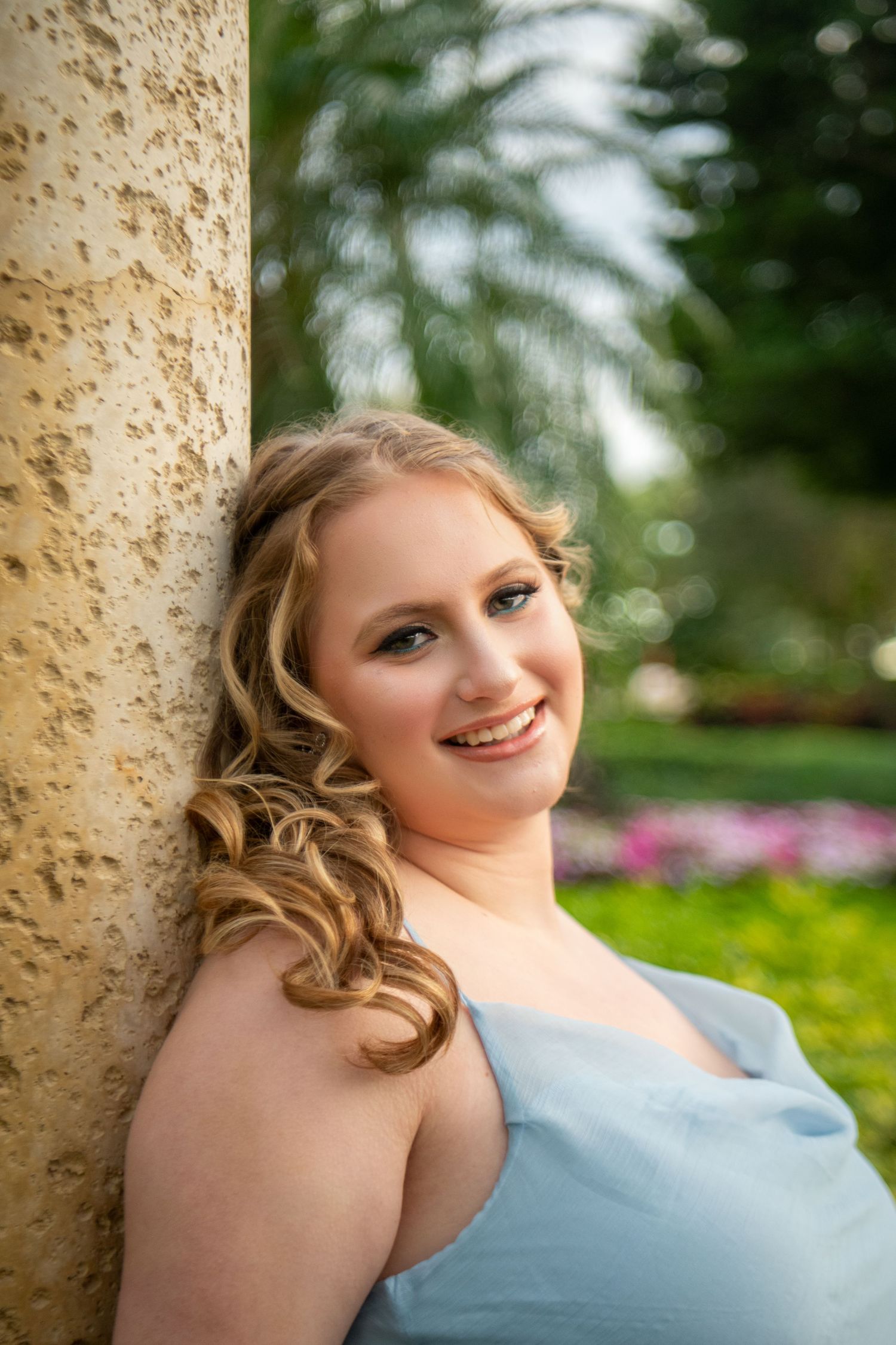 Portrait in soft blue dress against stone pillar with blurred garden background and natural lighting.