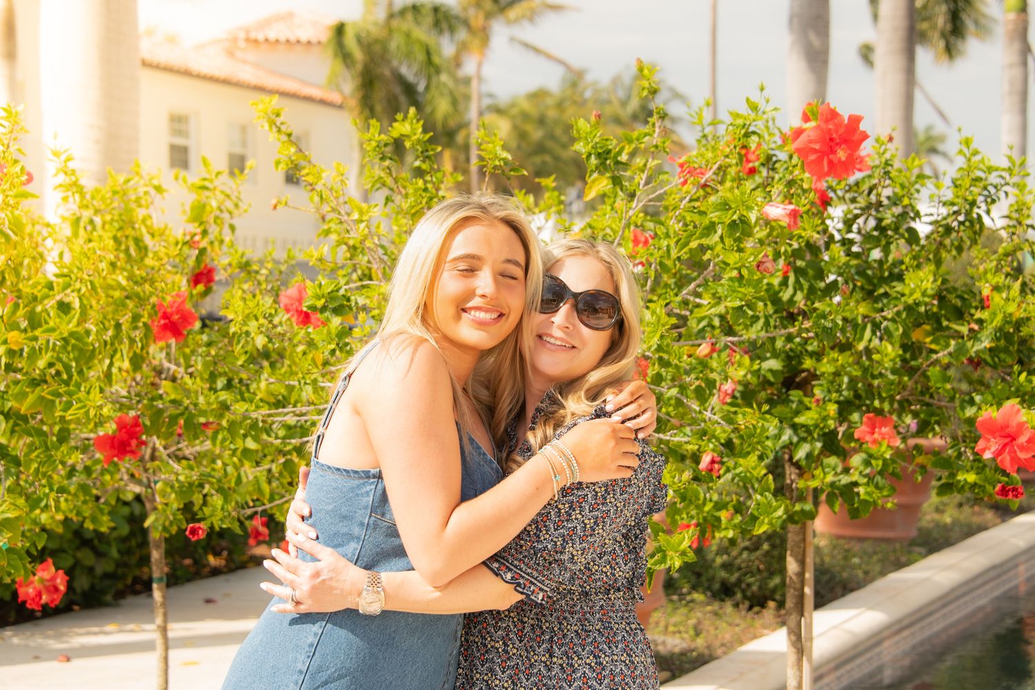 Two people embrace and smile in a sunny garden setting with vibrant red flowers and palm trees in the background.