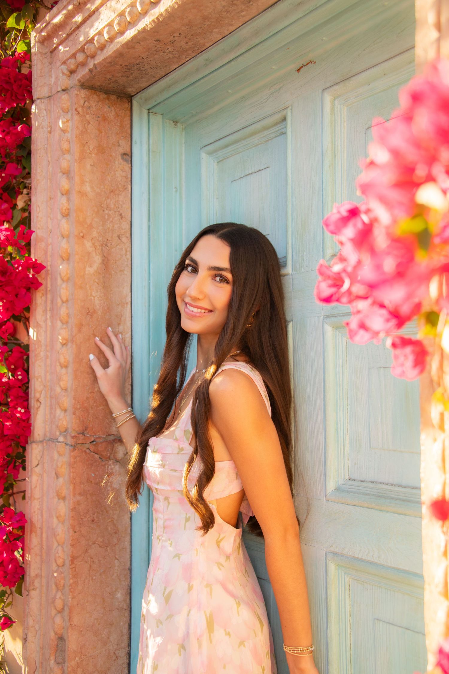 A person in a pink dress poses by a turquoise door framed with vibrant pink bougainvillea flowers.