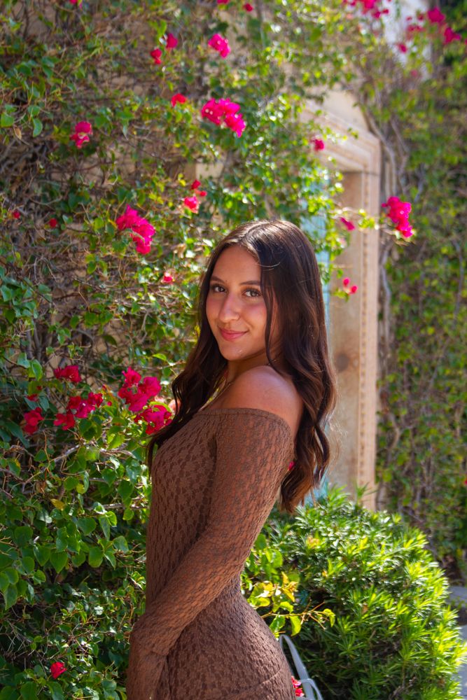 A woman in a brown knit sweater poses gracefully in a garden with blooming red roses and lush greenery.