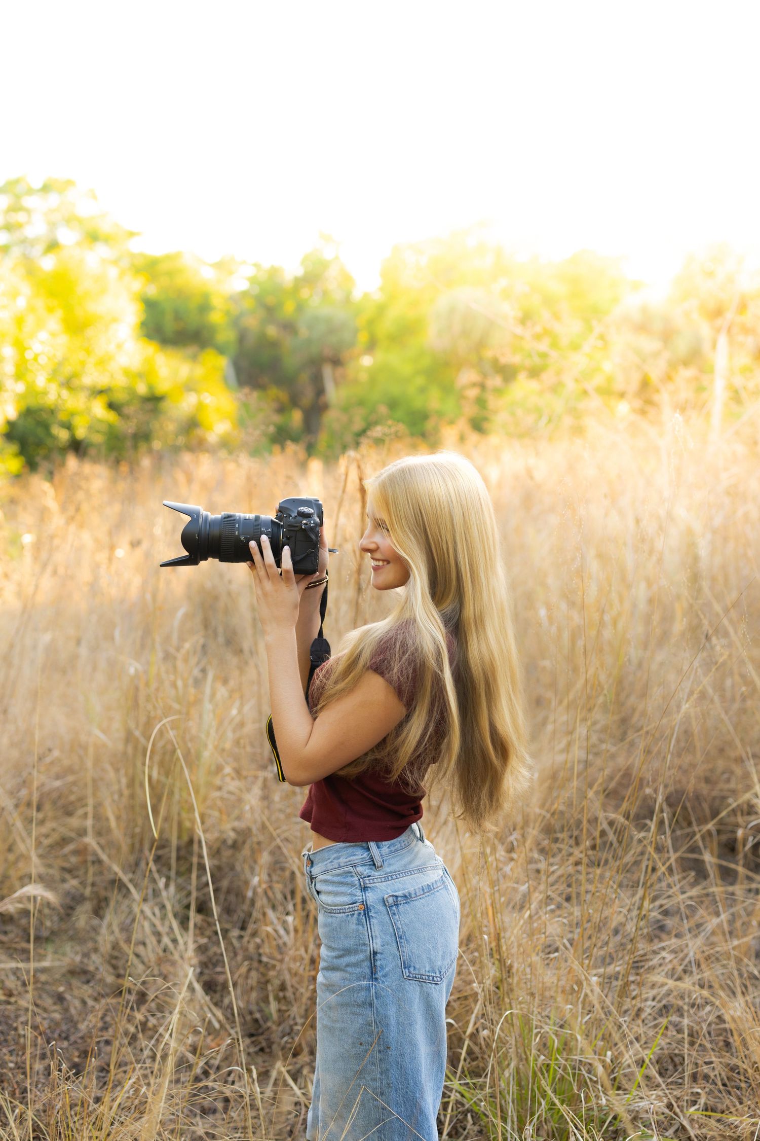 Photographer in burgundy top and jeans captures nature shots in golden field during sunset.