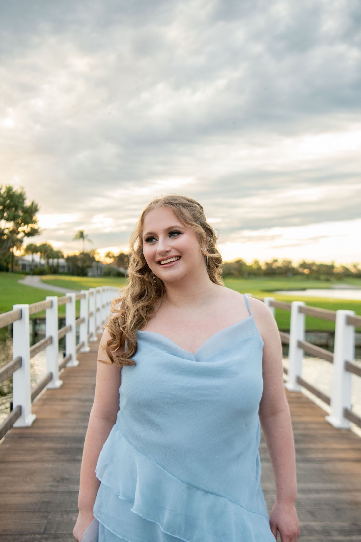 Woman in light blue dress poses on wooden boardwalk at sunset with cloudy sky and water in background.