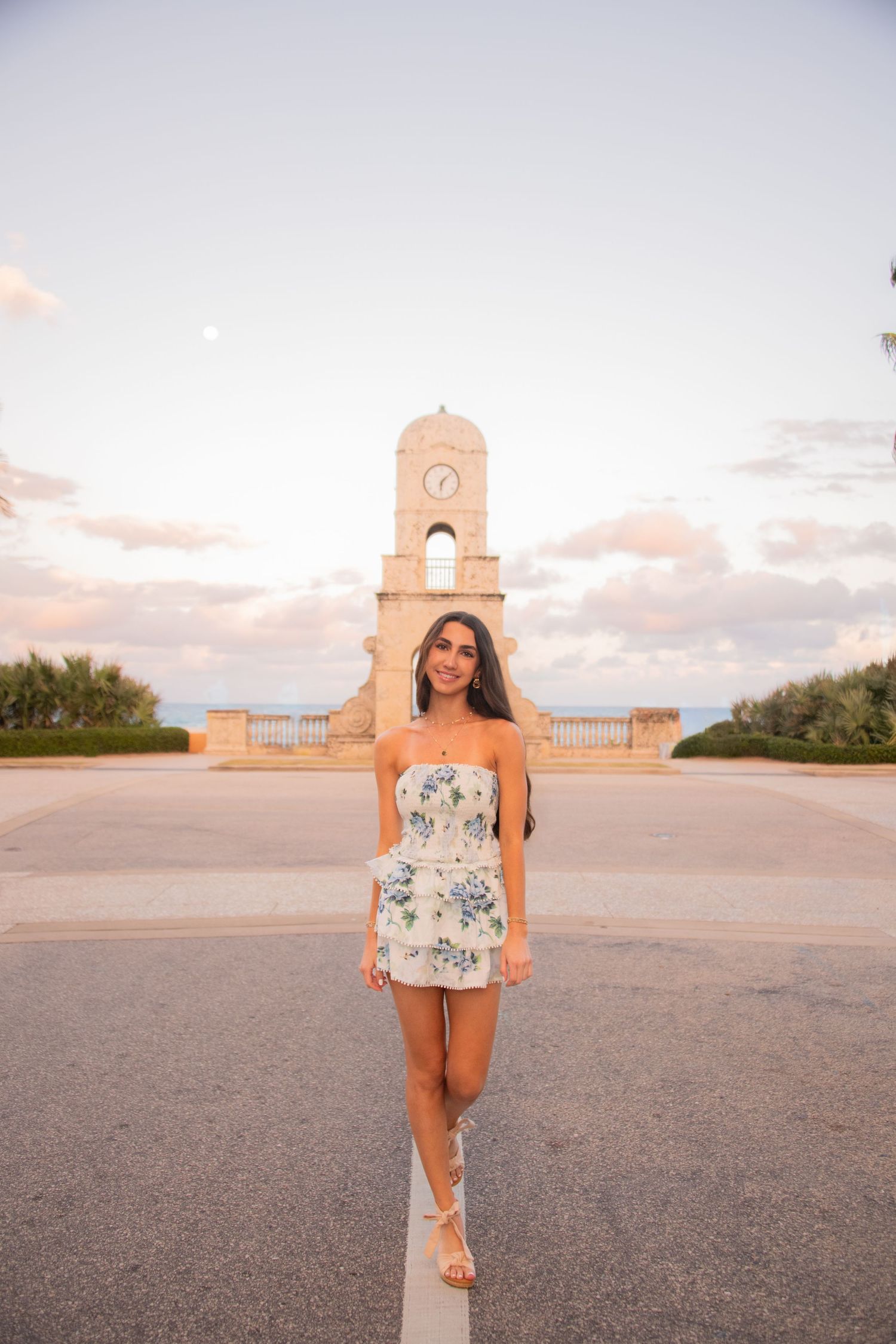 A person poses in a floral romper at sunset with a stone monument and cloudy sky in the background.