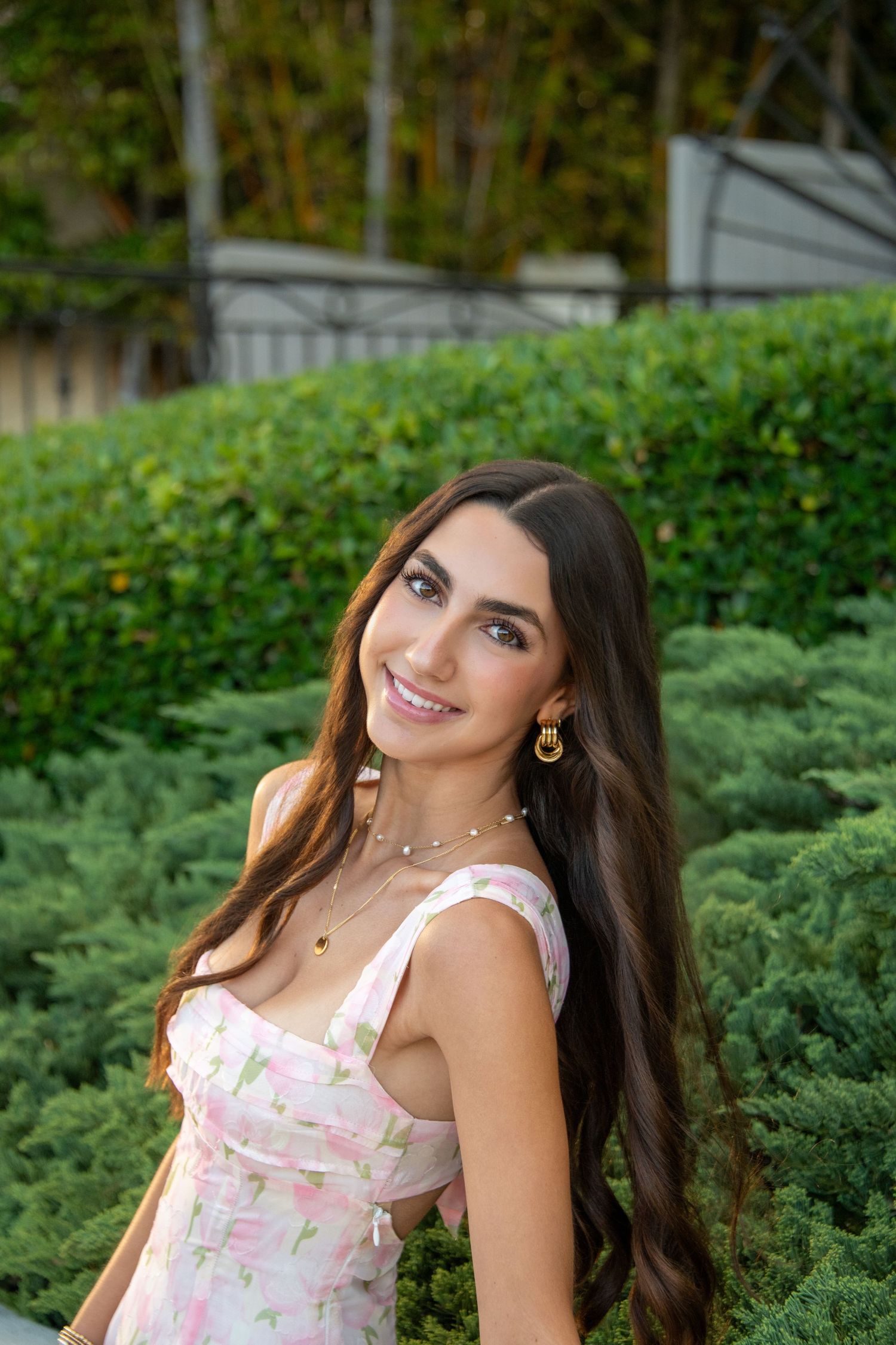A person in a floral dress poses outdoors against lush green foliage in a garden setting.