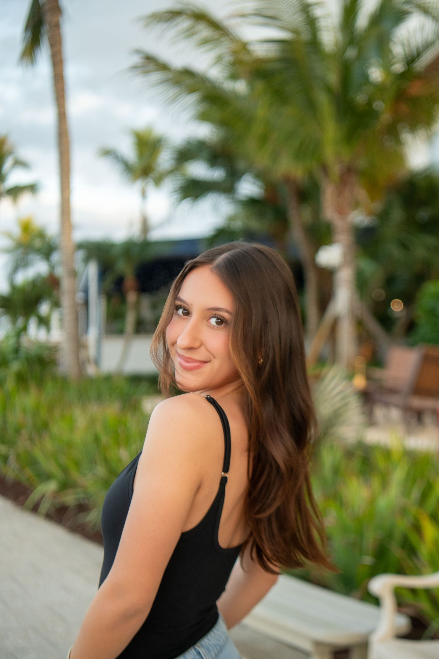 Young woman in black tank top poses outdoors against tropical palm trees and beach backdrop during sunset.