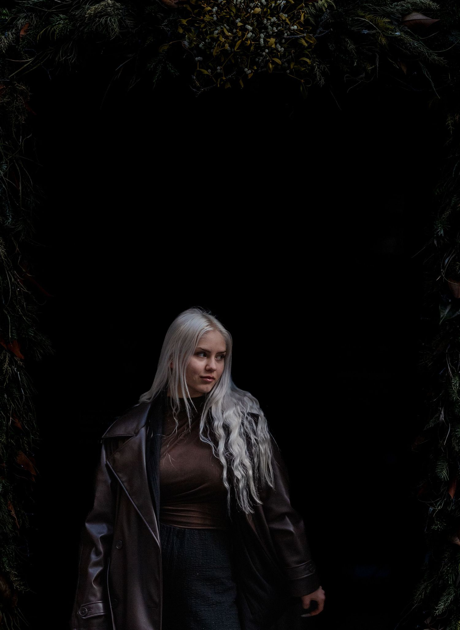 Moody portrait against dark foliage backdrop showing figure in leather jacket with flowing white hair.