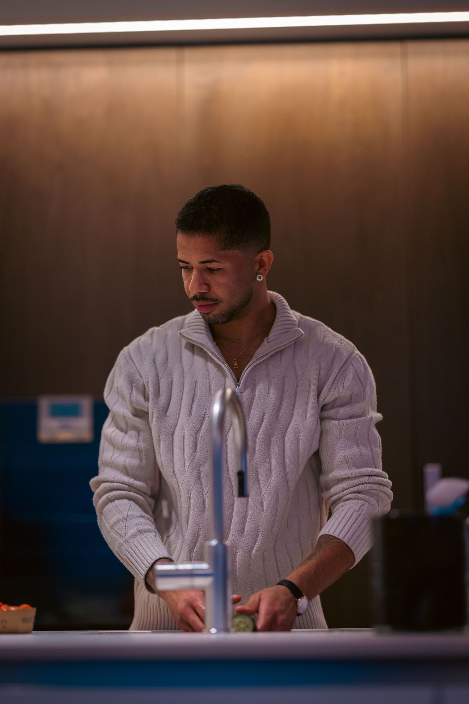 A stylish man in a white cable-knit sweater stands at a kitchen counter in moody ambient lighting.