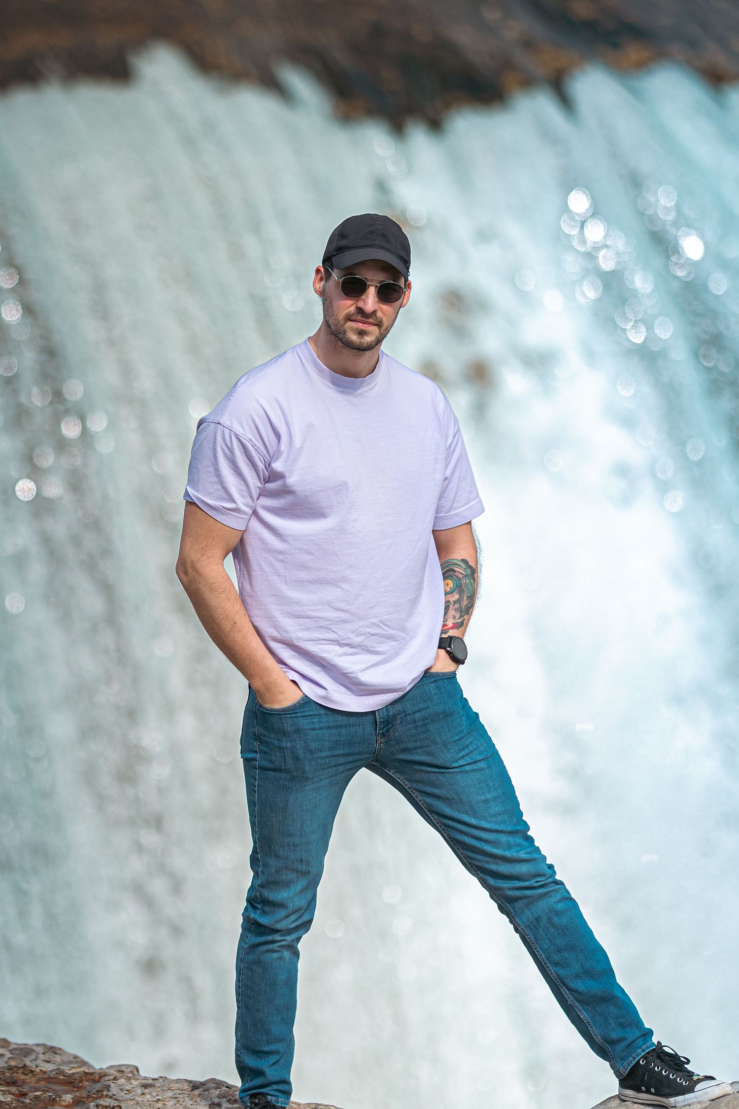 Man in white t-shirt, jeans, and cap poses confidently in front of a cascading waterfall outdoors.