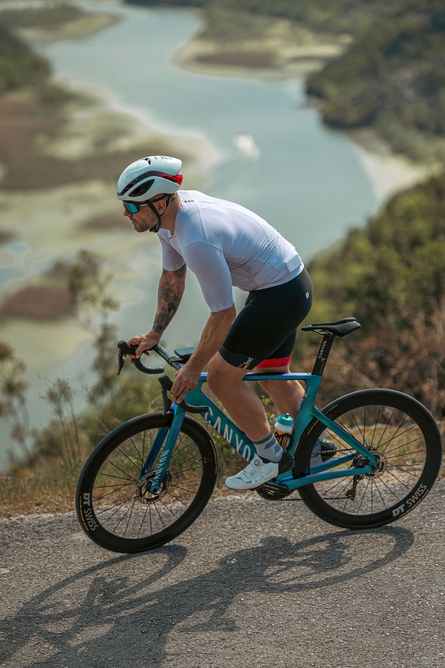 Cyclist in white jersey rides teal Canyon bike on mountain road with stunning turquoise river valley below.