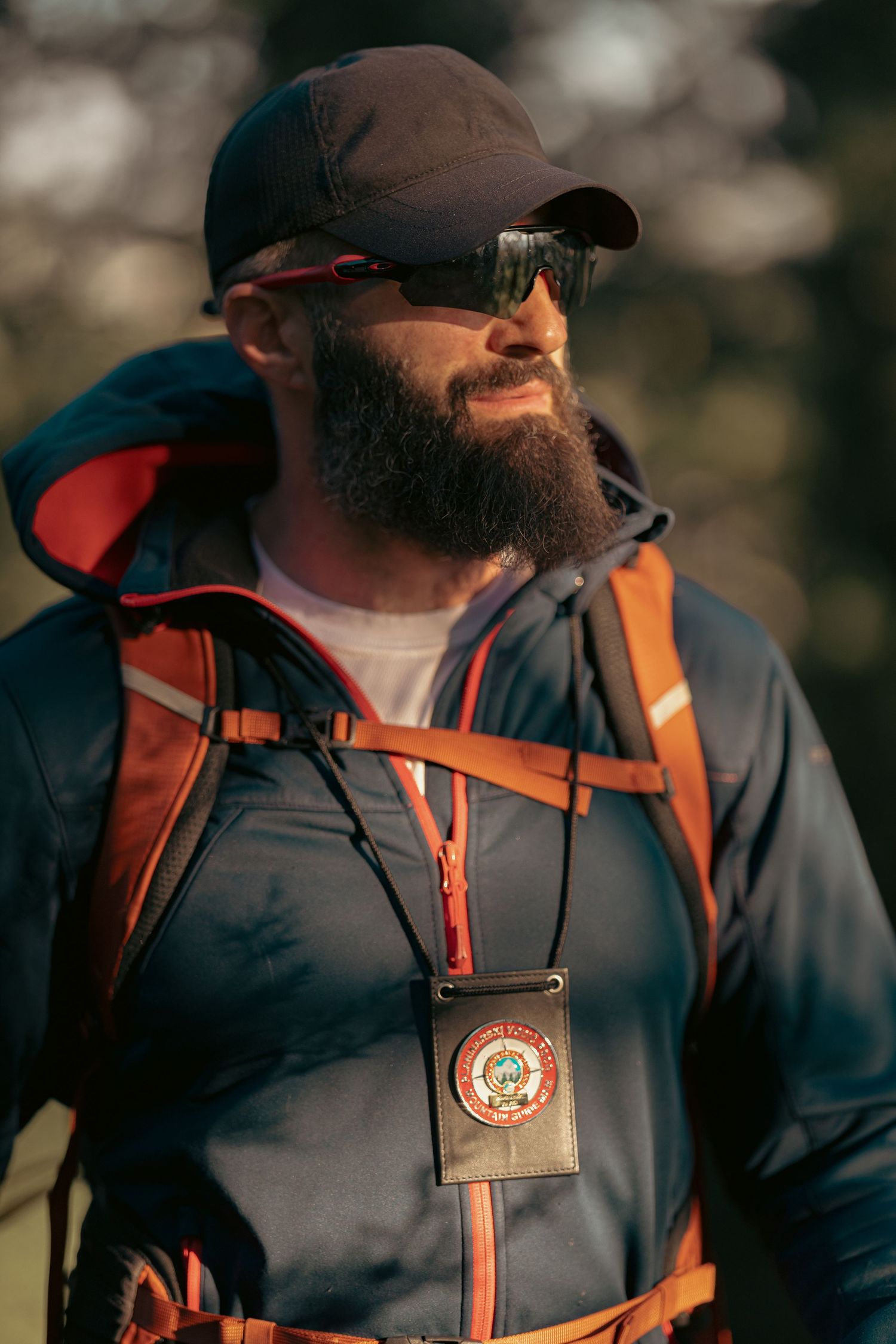 Bearded male hiker wearing sunglasses, cap, and backpack with badge outdoors in natural light.