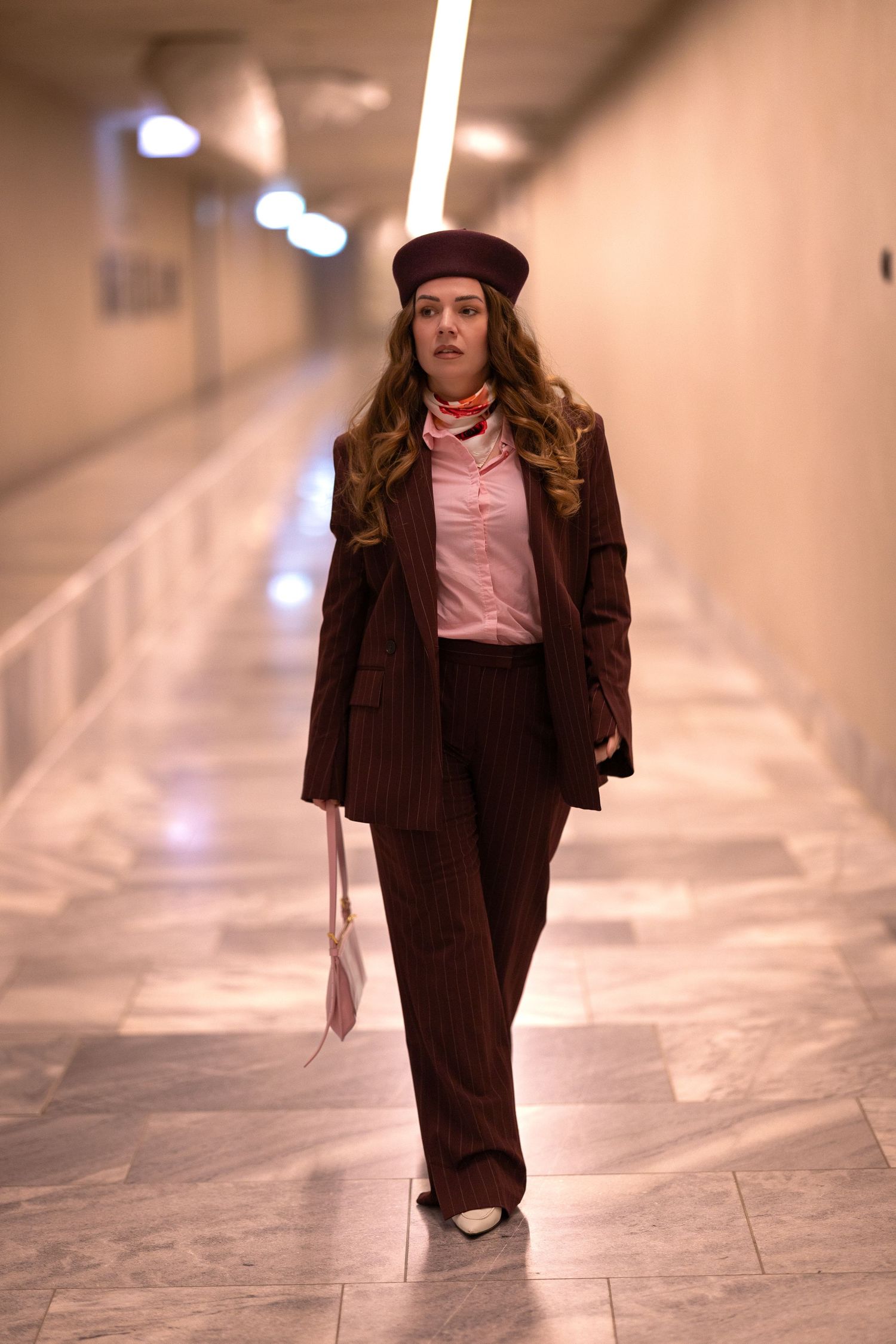 A person wearing a brown suit and beret walks through a bright indoor space with tiled floors.
