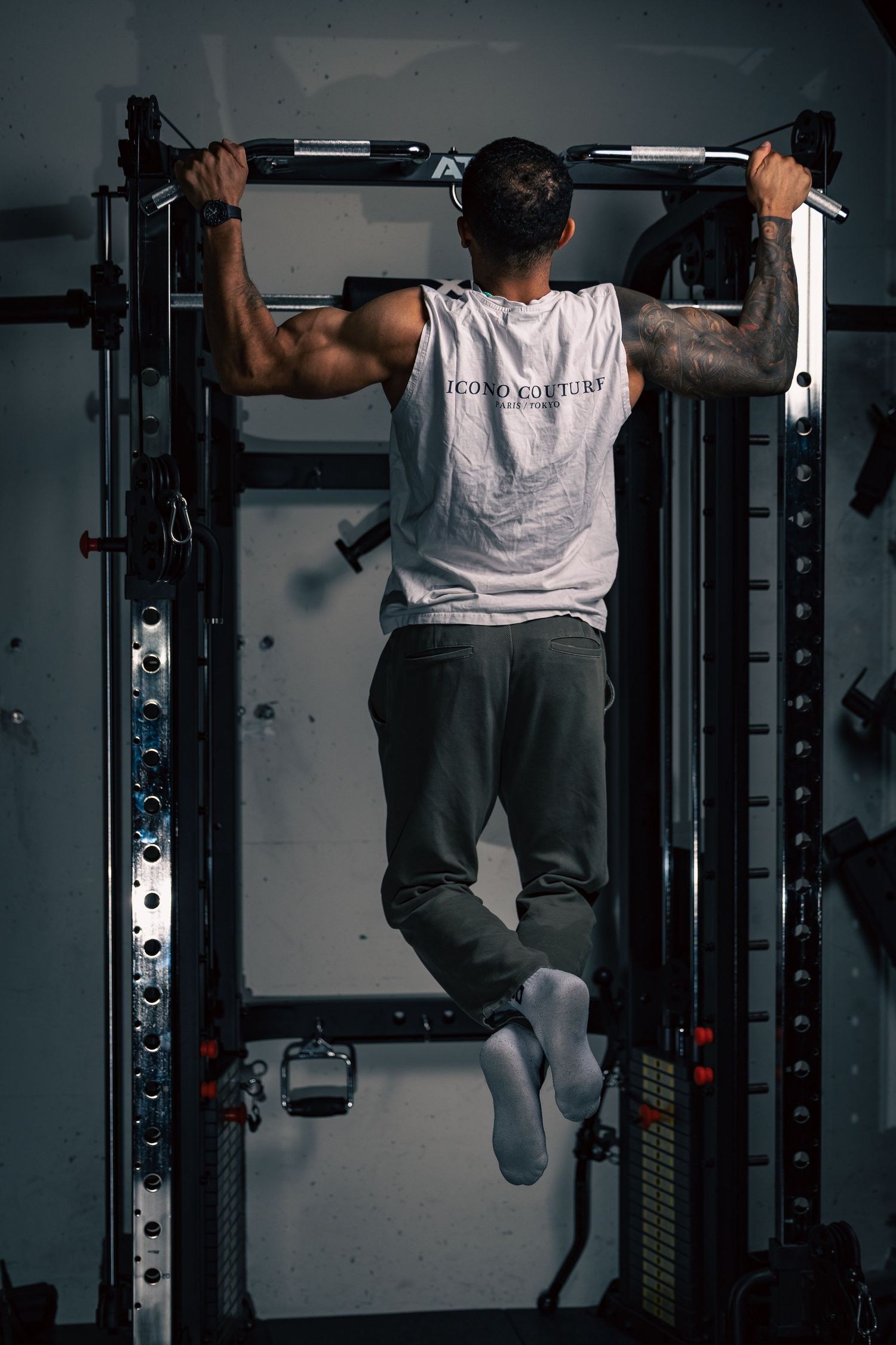 Man performing pull-ups on a power rack in a gym, wearing a white tank top and gray pants, showing back muscles.