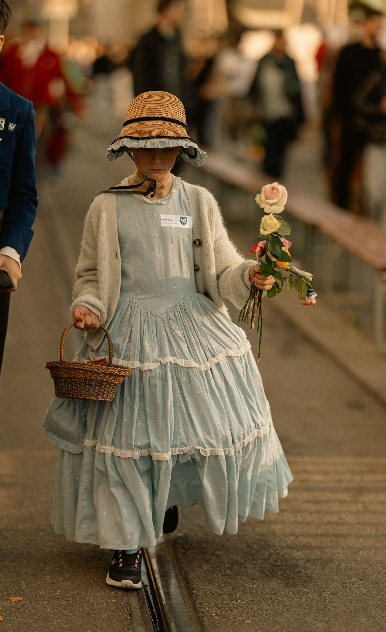 A young girl in a light blue ruffled dress, straw hat, and cardigan carries a basket and bouquet of roses.
