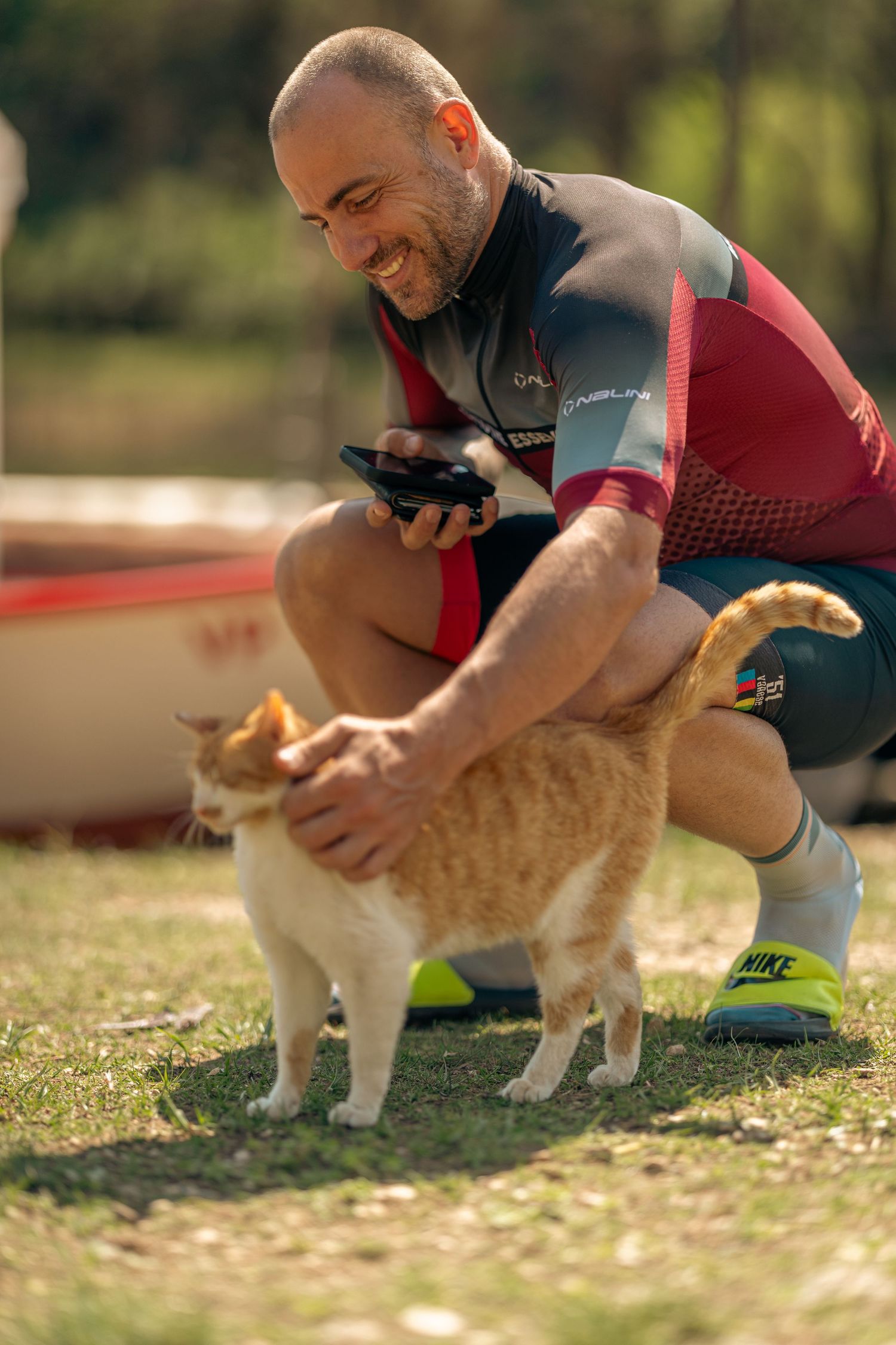 Smiling bald man in cycling jersey crouching to pet an orange tabby cat outdoors near a boat.
