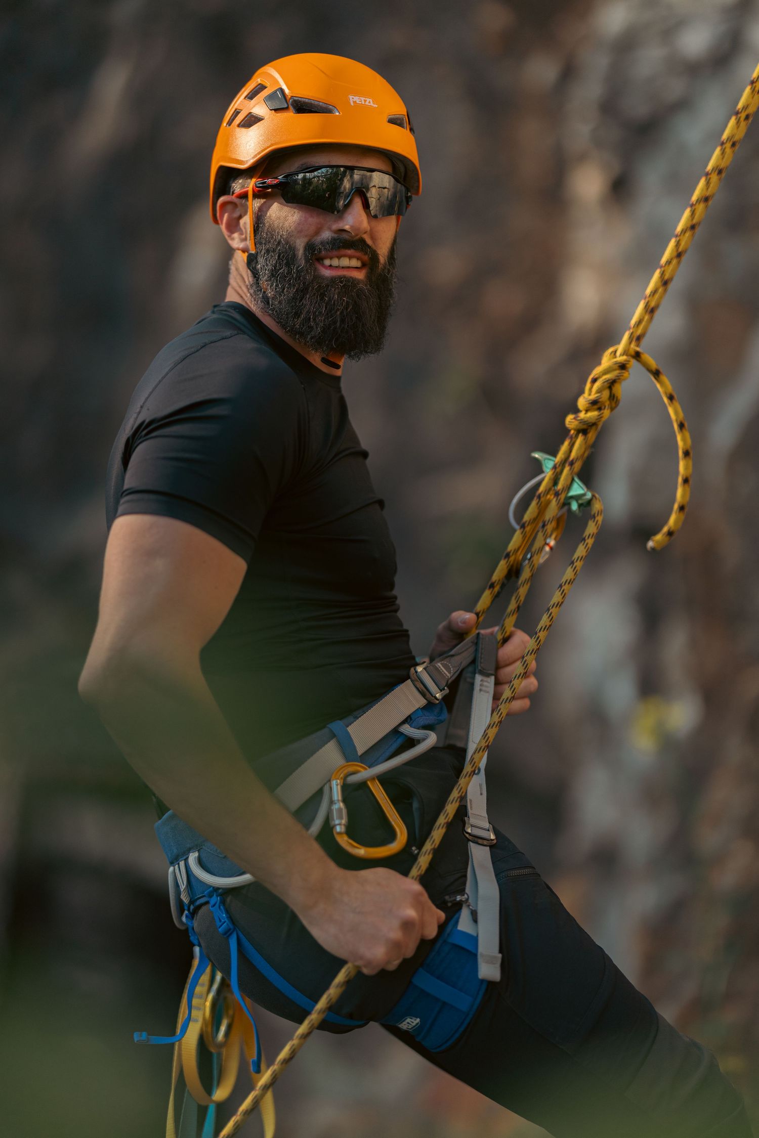Bearded male rock climber wearing orange helmet and sunglasses with safety harness and ropes on a cliff face.