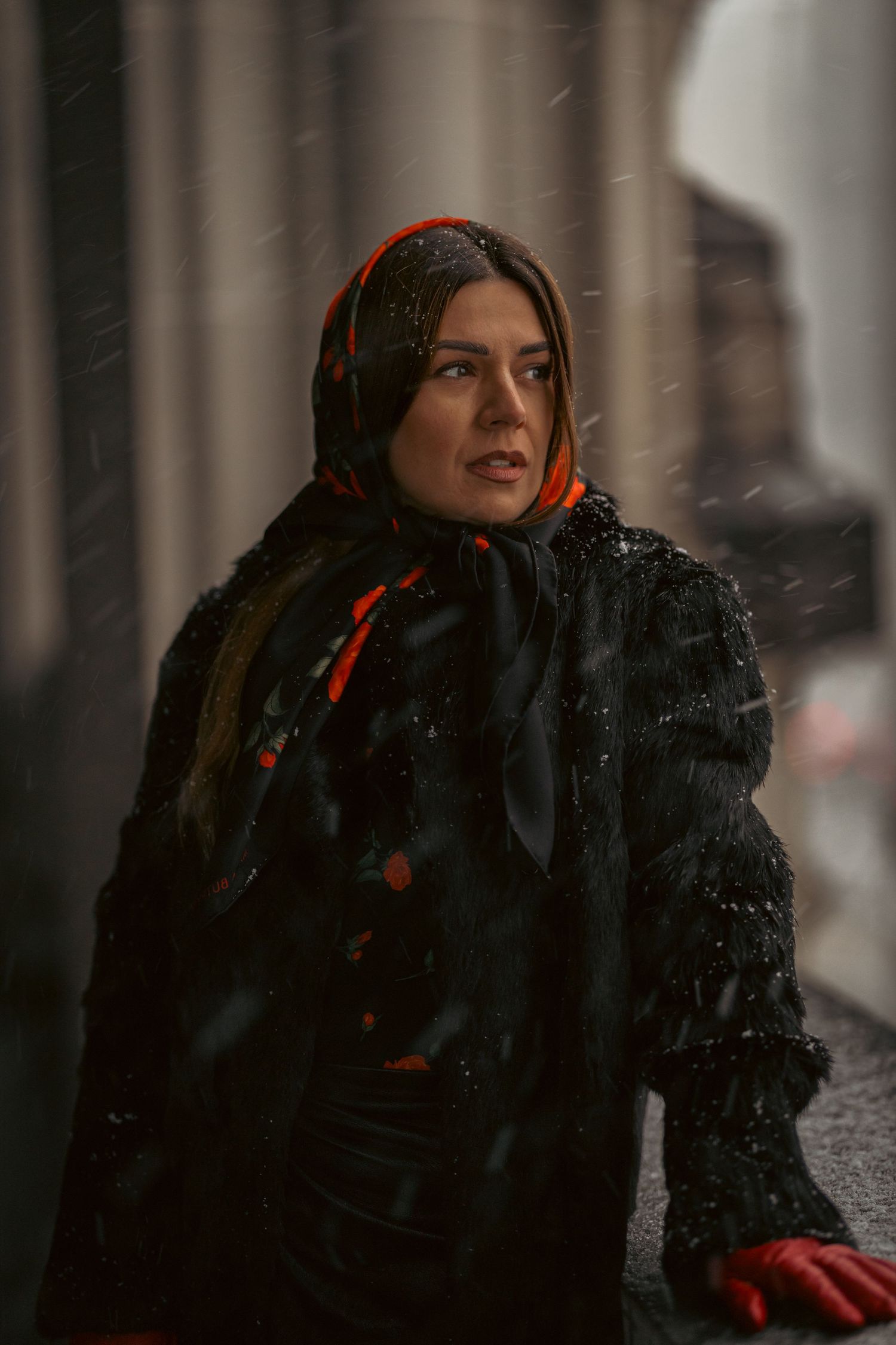 A person in a dark winter coat with a red scarf looks up at falling snow on a gloomy winter day.