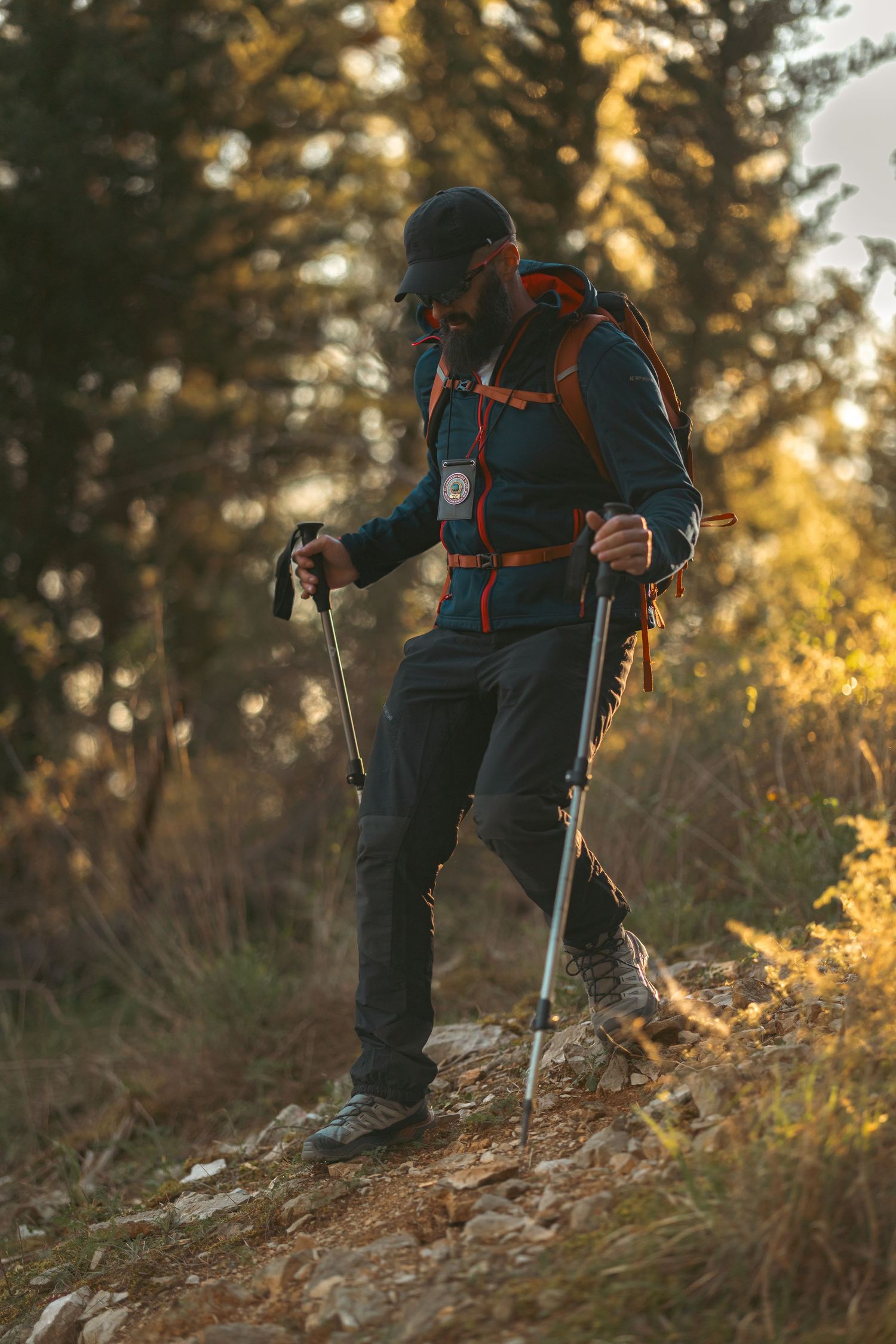 Hiker with trekking poles and orange backpack navigating rocky mountain trail through autumn forest at sunset.
